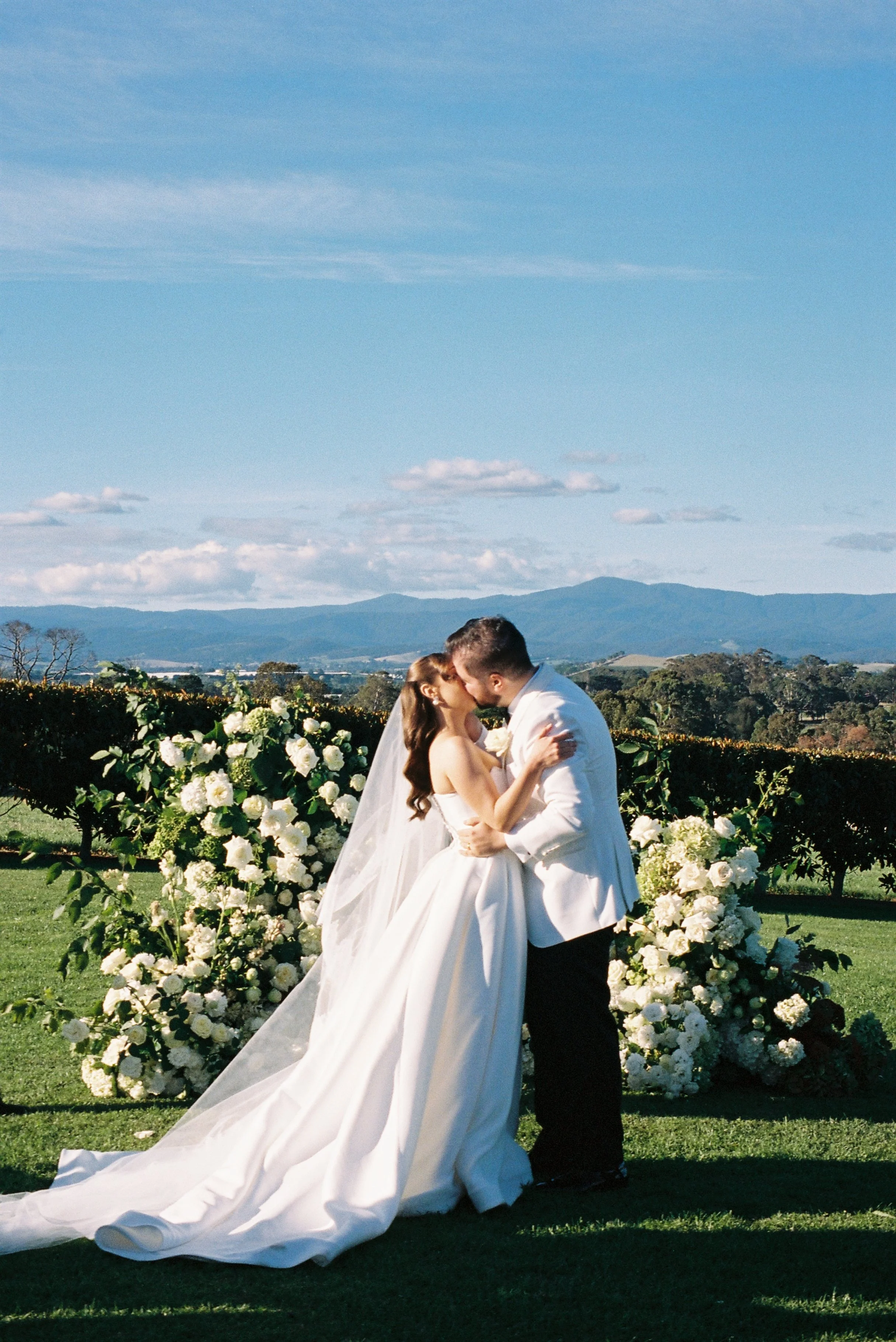 A bride and groom kissing during their outdoor wedding ceremony, surrounded by white flowers and greenery, with a scenic mountain landscape in the background.