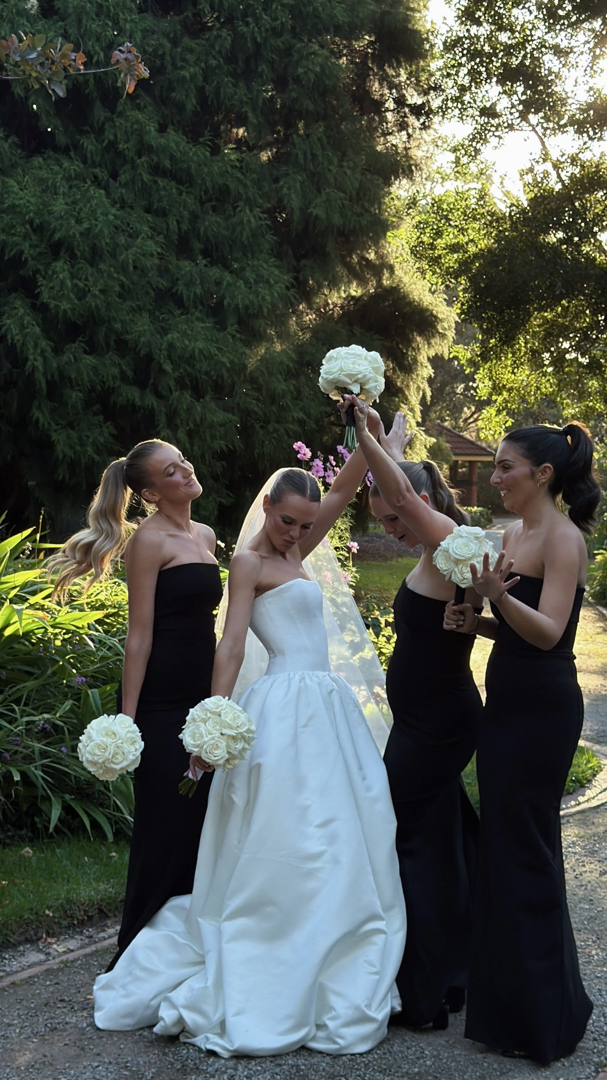 Four women, including a bride in a white wedding gown and three bridesmaids in black dresses, celebrating outdoors with bouquets of white roses on a sunny day with lush green trees in the background.