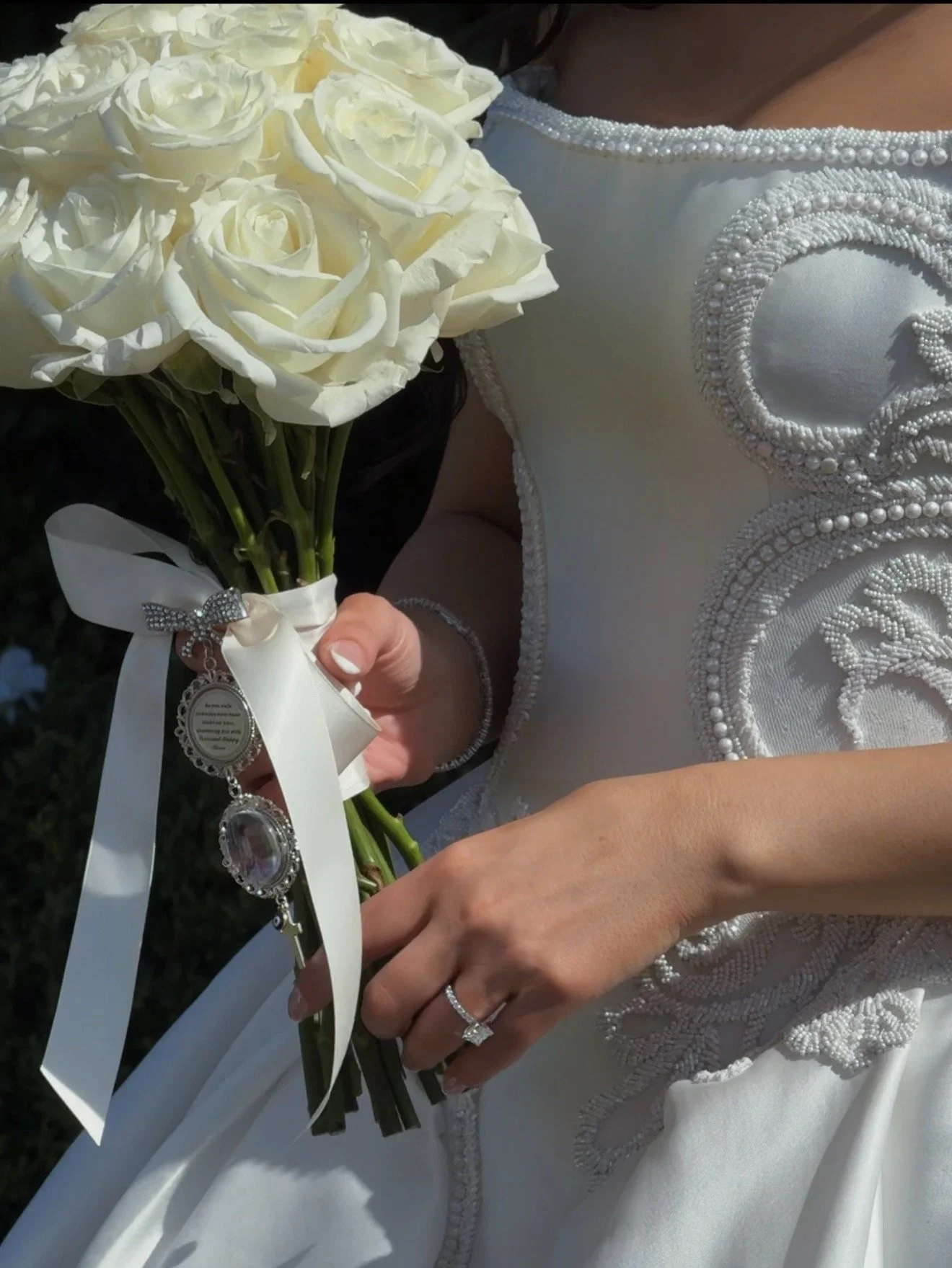 Close-up of a bride holding a bouquet of white roses, dressed in a wedding gown with intricate pearl and bead embellishments.