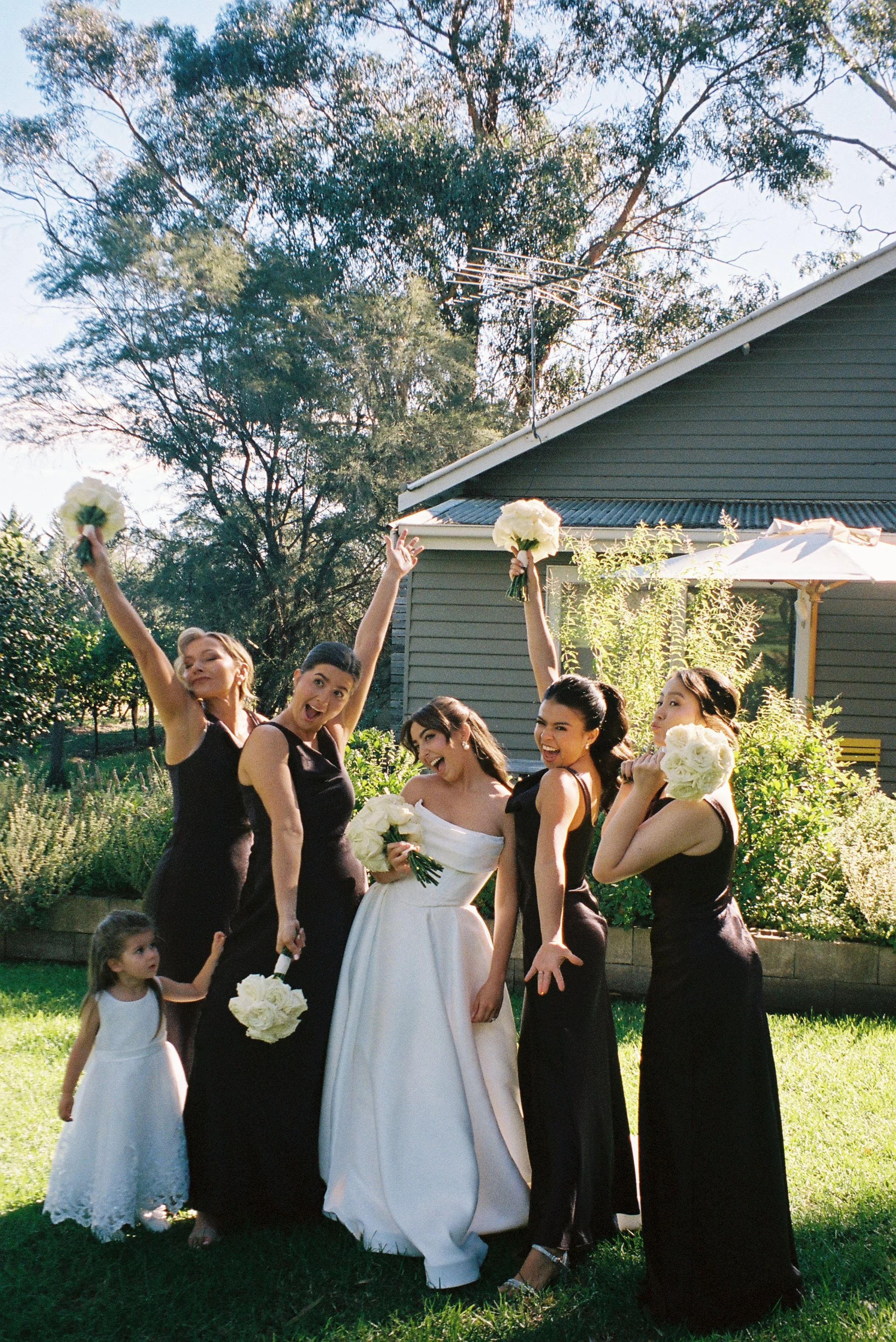 Group of women in formal dresses celebrating outdoors, some holding bouquets, with a house and trees in the background.