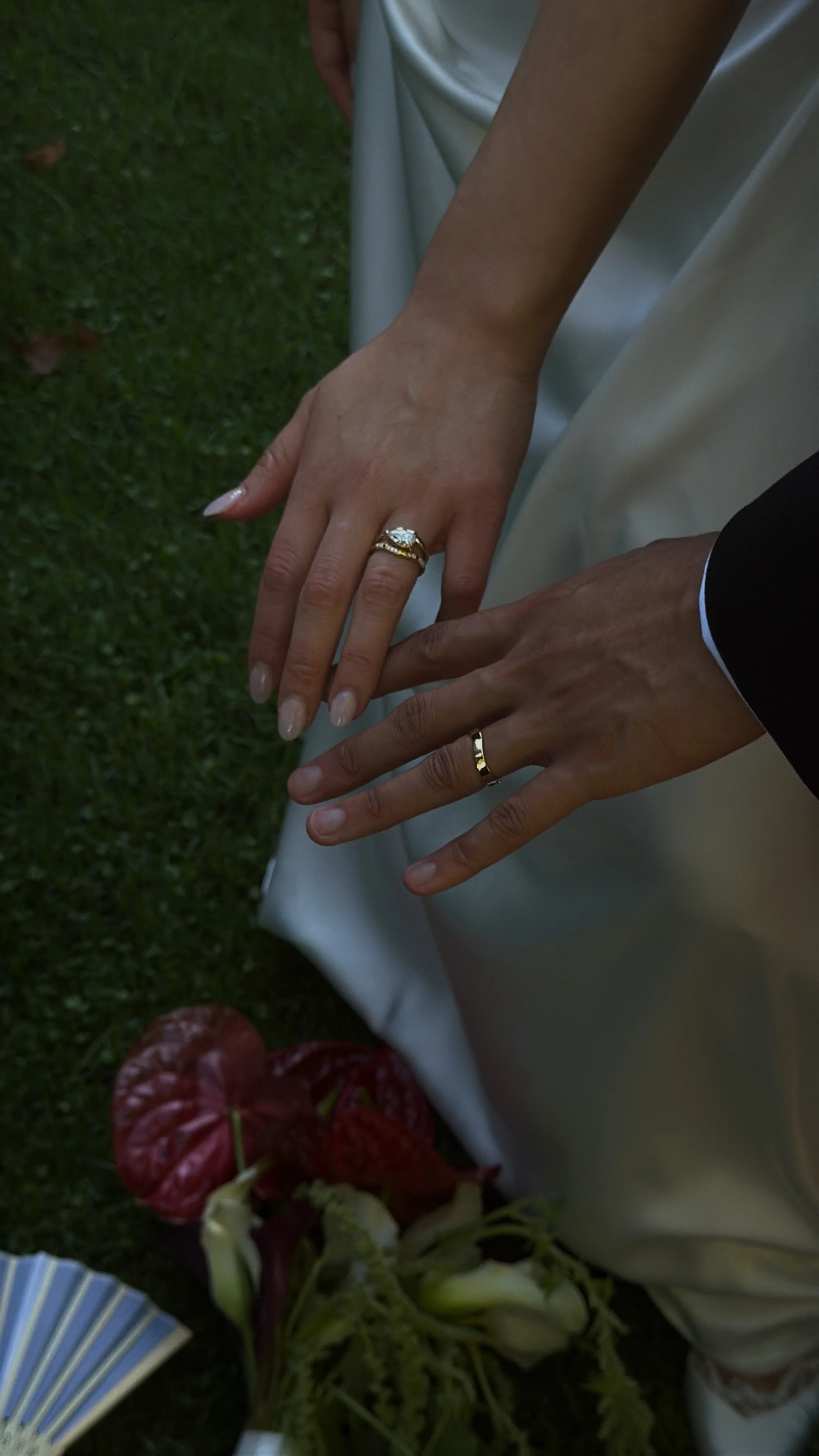 Close-up of a person's hands displaying wedding rings, with a wedding dress and green grass background.
