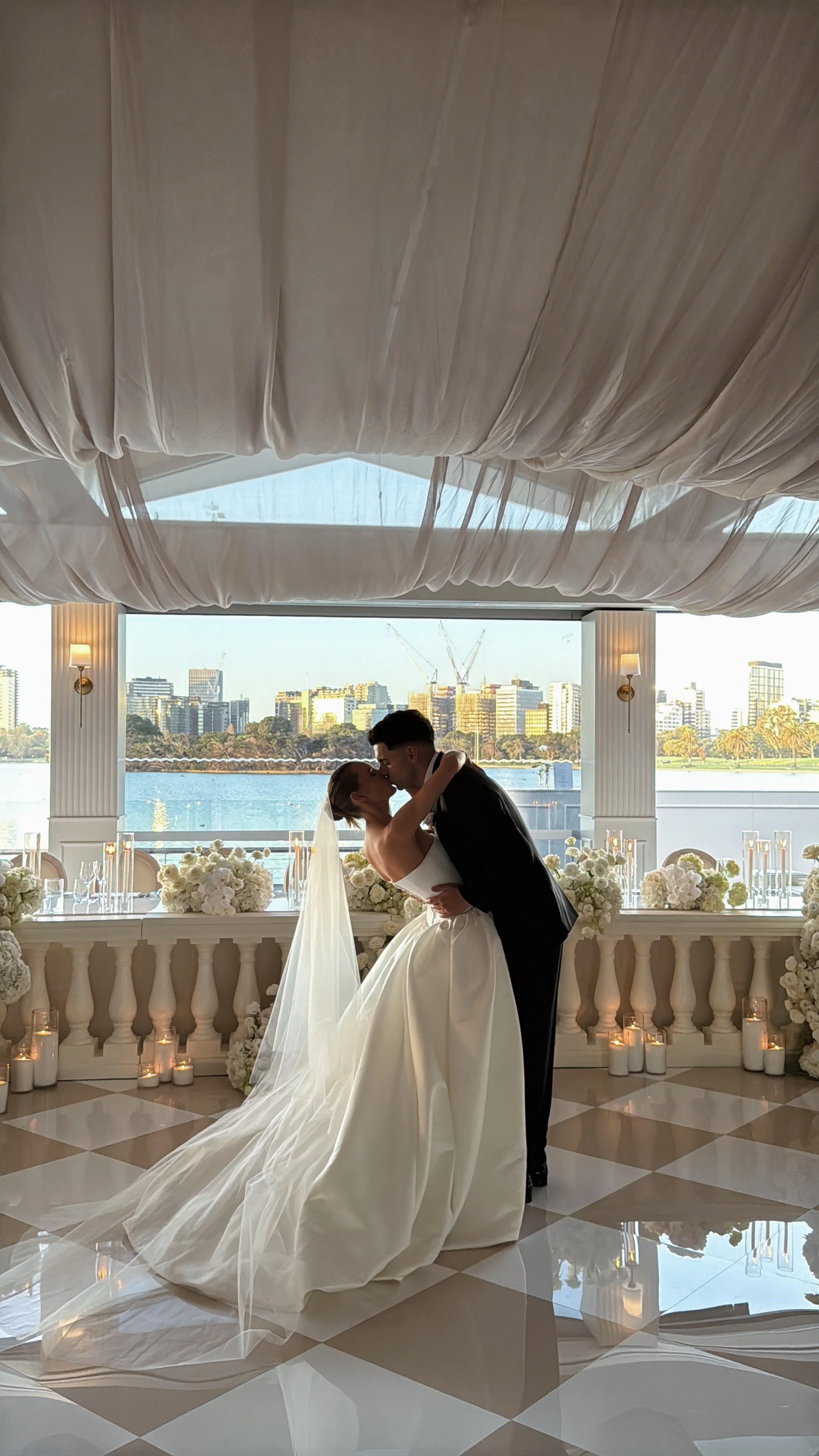 A bride and groom sharing a kiss during their wedding in a beautifully decorated indoor venue with large windows, overlooking a city skyline with river, modern buildings, floral arrangements and candles on the window ledge.