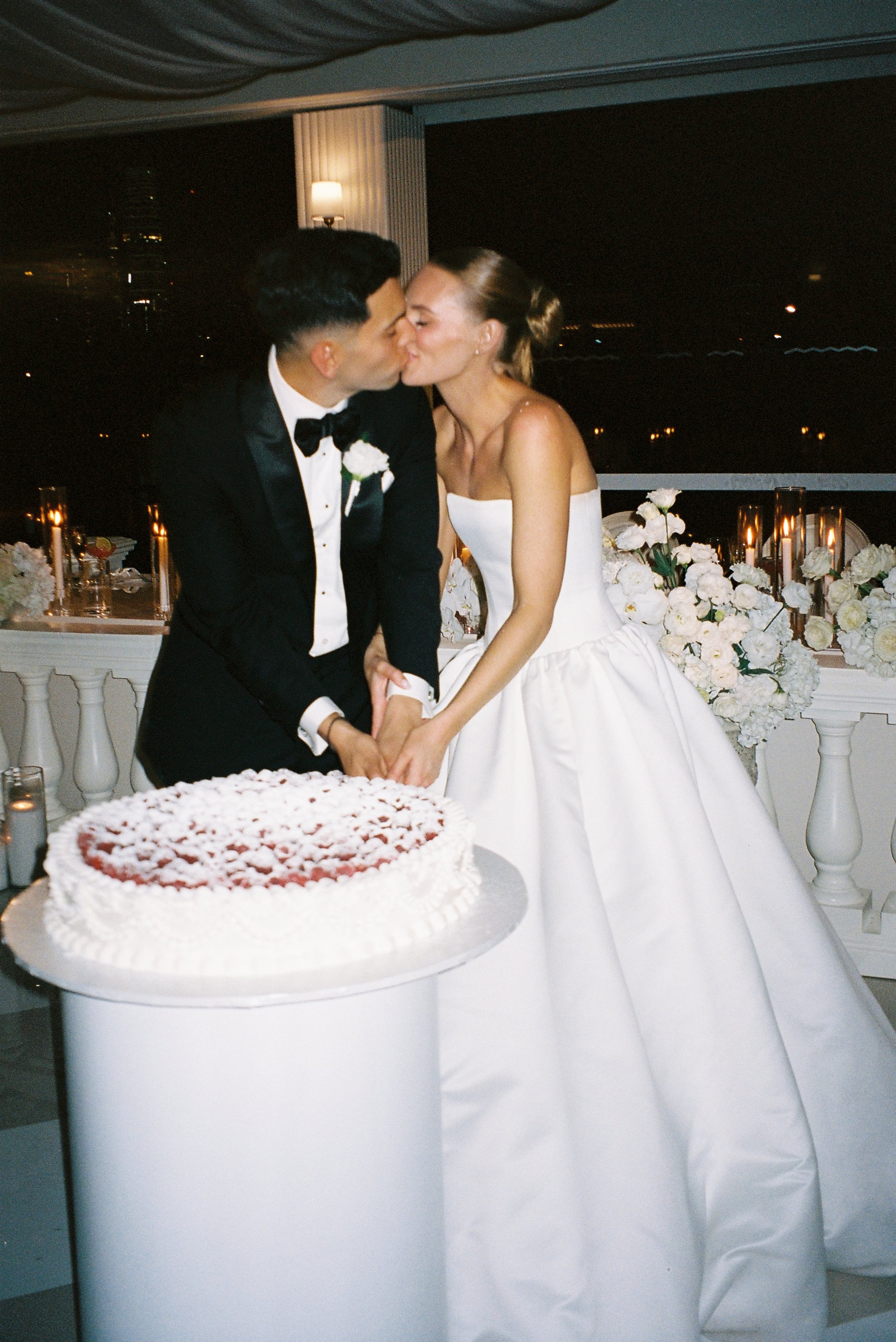 A bride and groom kiss at their wedding reception, with a cake on the table in front of them and a night city view in the background.
