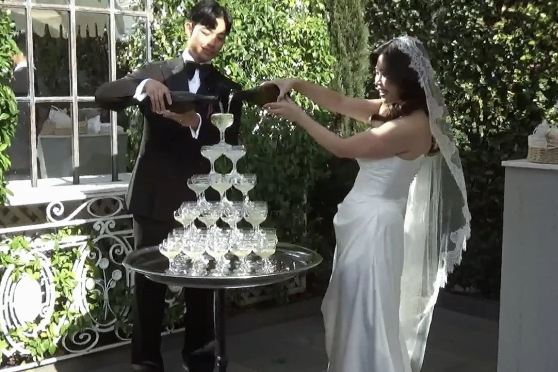 Bride and groom in wedding attire at an outdoor reception, pouring champagne into a pyramid of glasses.