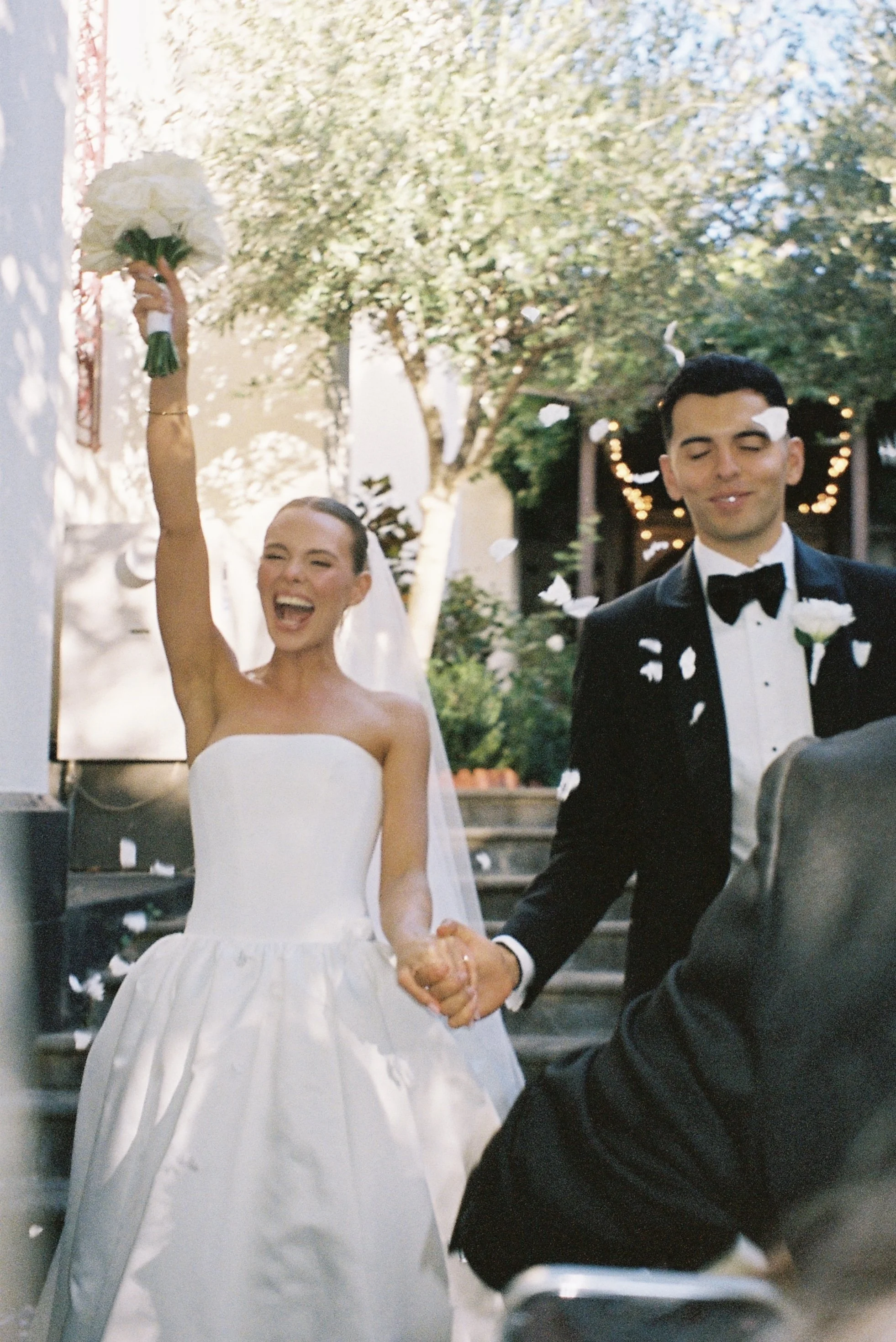 A bride and groom are holding hands and smiling, with the bride raising a bouquet in celebration and the groom wearing a tuxedo, during a wedding outside under trees with sunlight and falling flower petals.