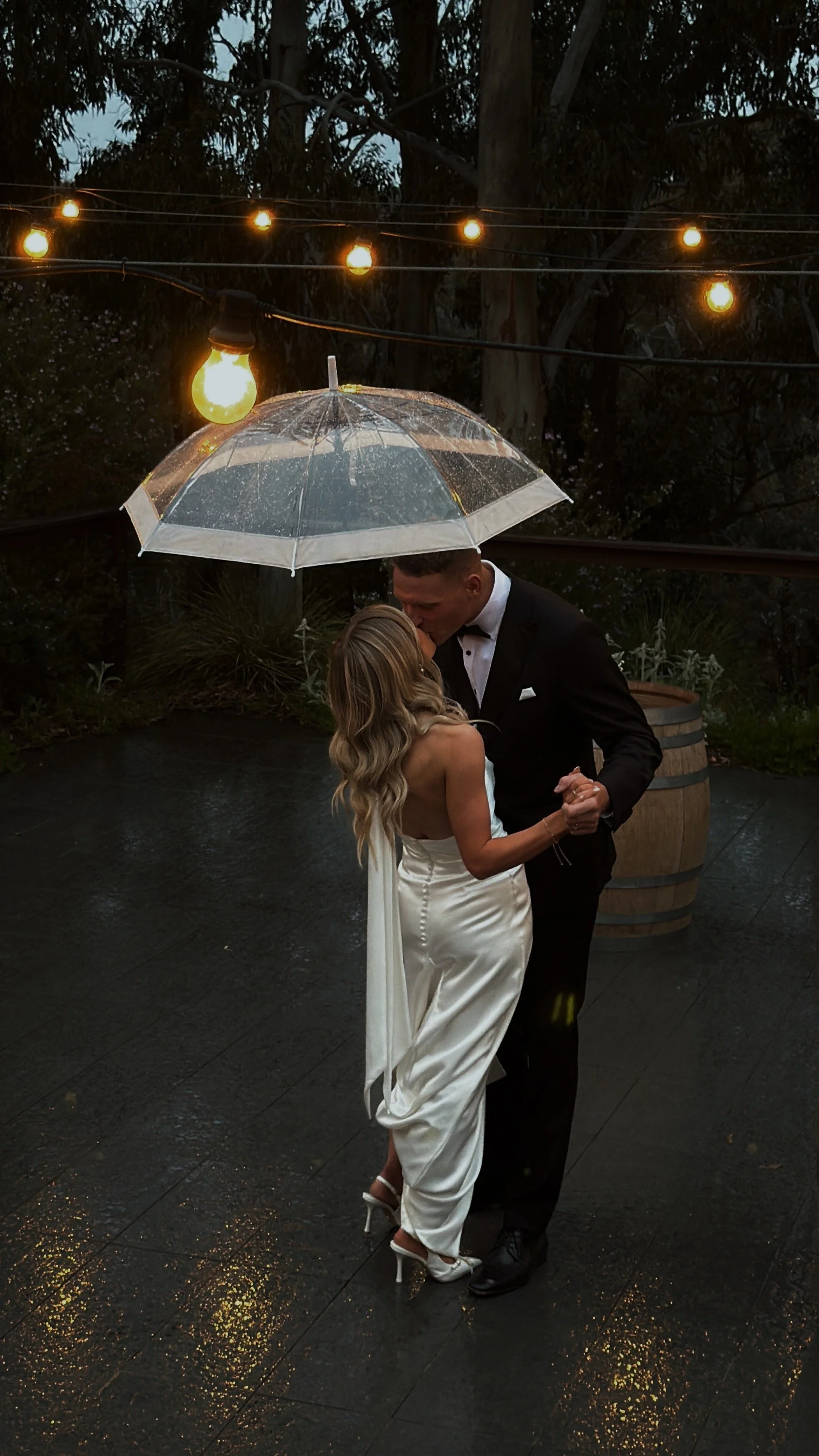 A bride and groom dancing under an umbrella on a rainy evening, with warm string lights overhead and a wet black pavement reflecting the lights.