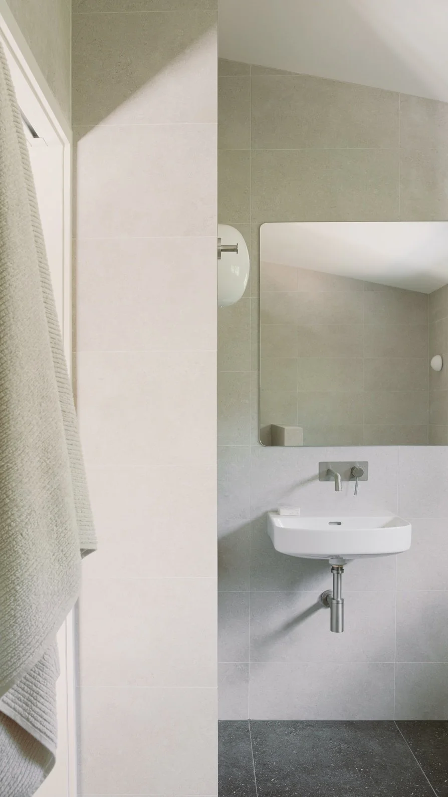A minimalist bathroom with a small white wall-mounted sink, a large mirror, gray tiled walls, and a towel hanging from a rack on the left side.