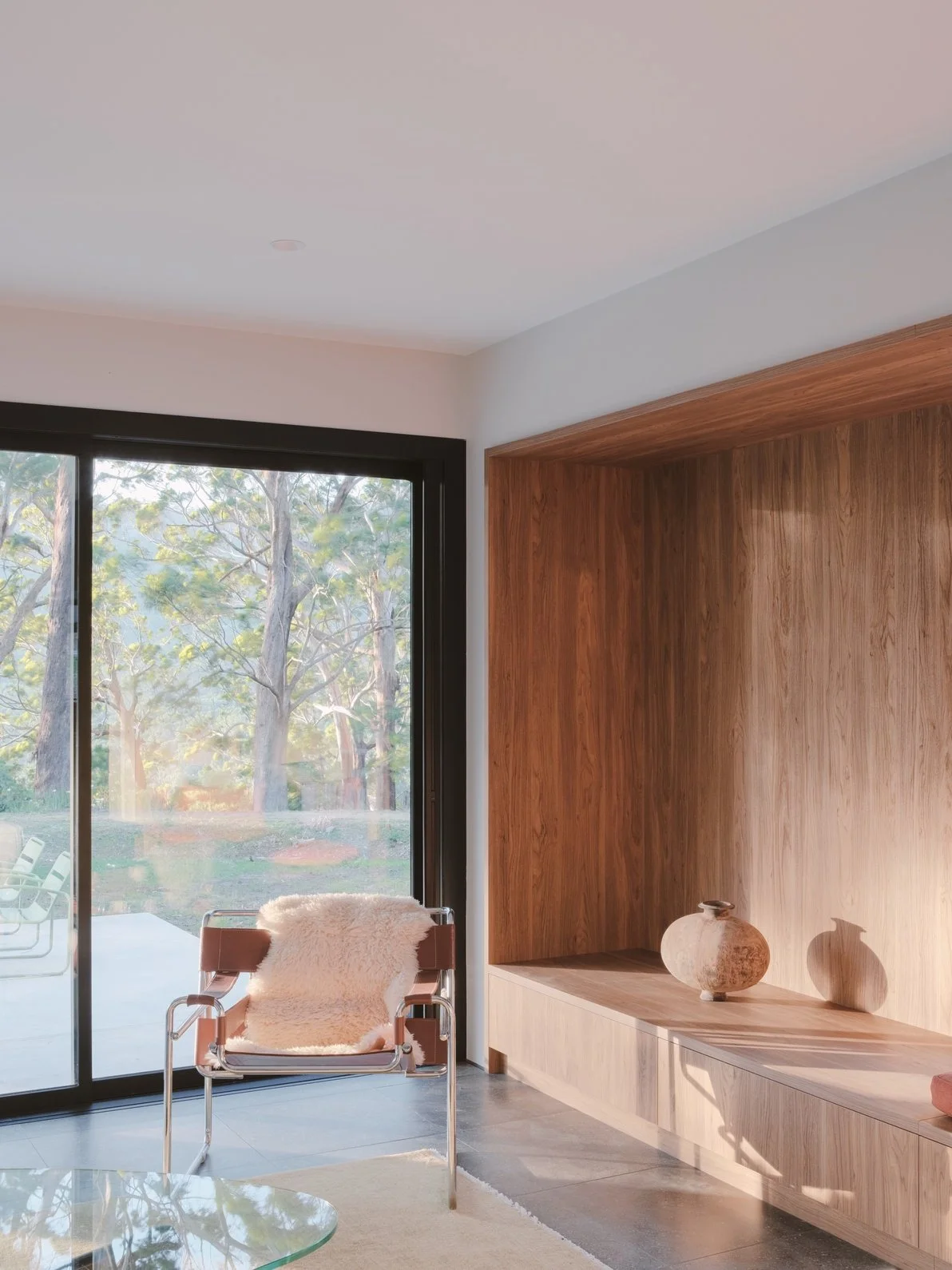 Interior of a modern living space with a large glass sliding door, a wooden wall feature with decorative vases, a leather chair with a sheepskin throw, and a view of a wooded outdoor area.