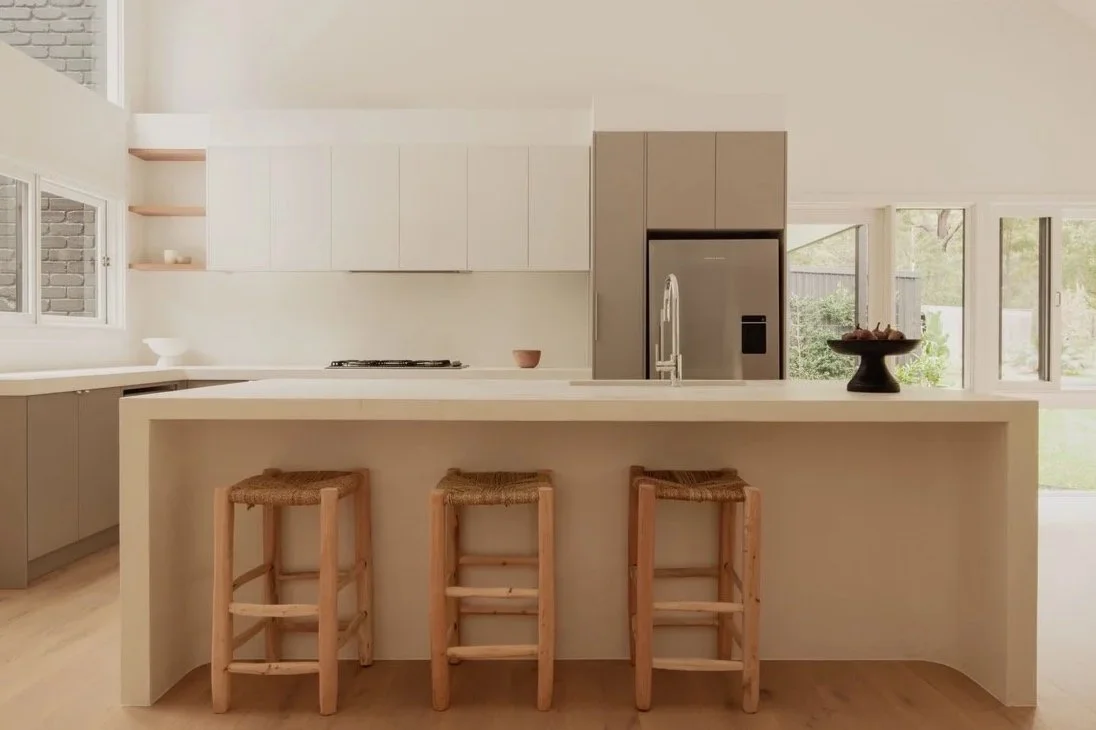 Modern kitchen with white and taupe cabinets, three wooden barstools at a white island, open shelves with minimal decor, stainless steel refrigerator, and large windows letting in natural light.