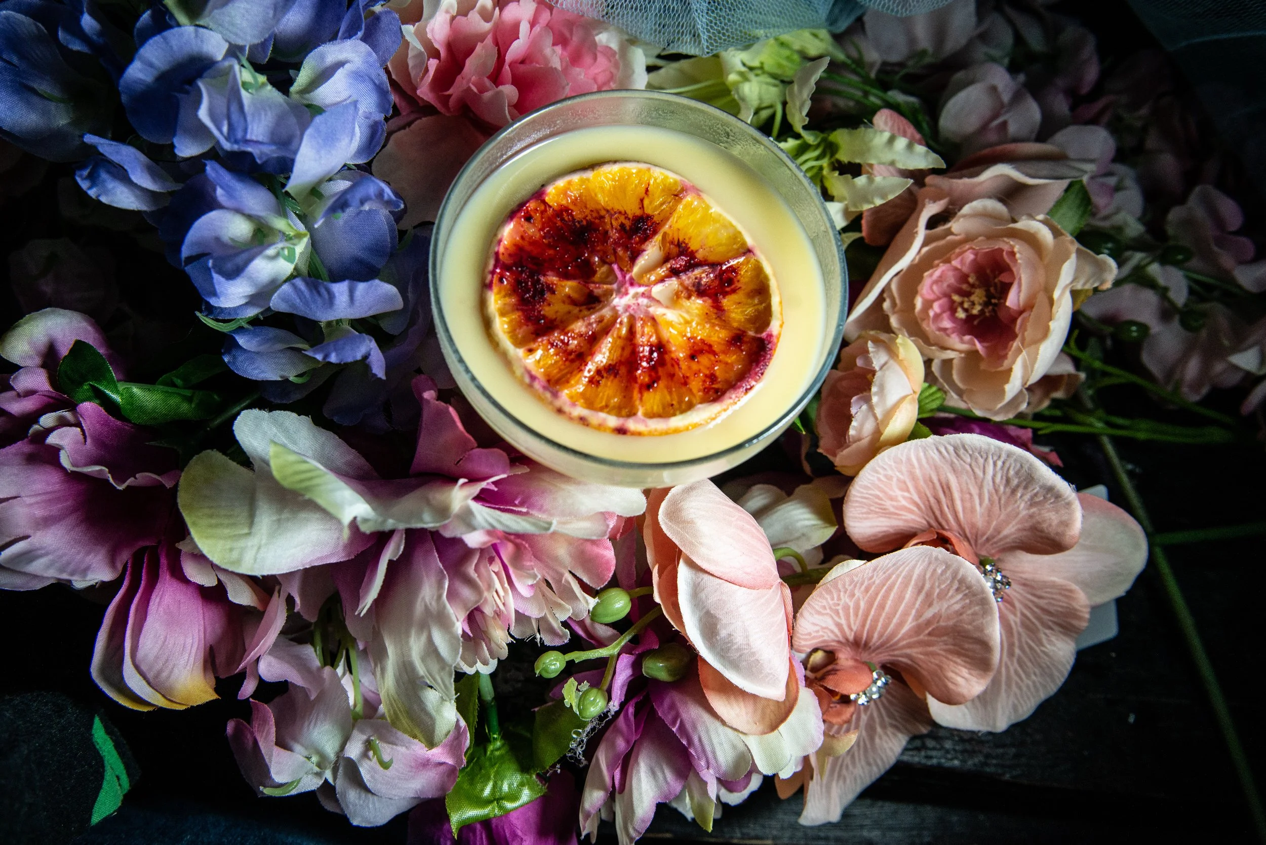 A glass of creamy dessert topped with a slice of orange, surrounded by pink, purple, and cream-colored artificial flowers.