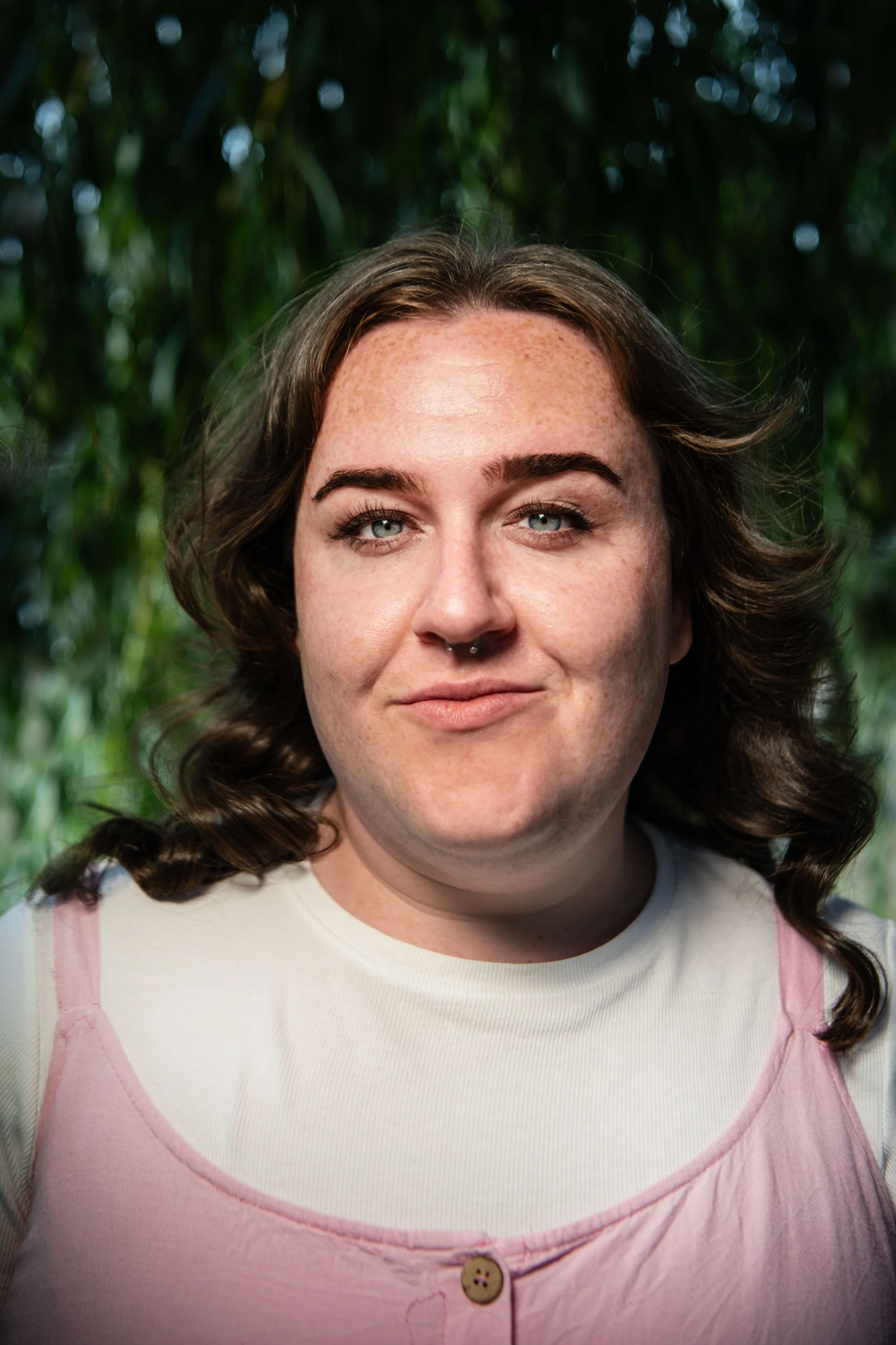 Close-up of a woman with light skin, freckles, wavy brown hair, blue eyes, and a septum piercing, standing outdoors with blurred green foliage in the background.