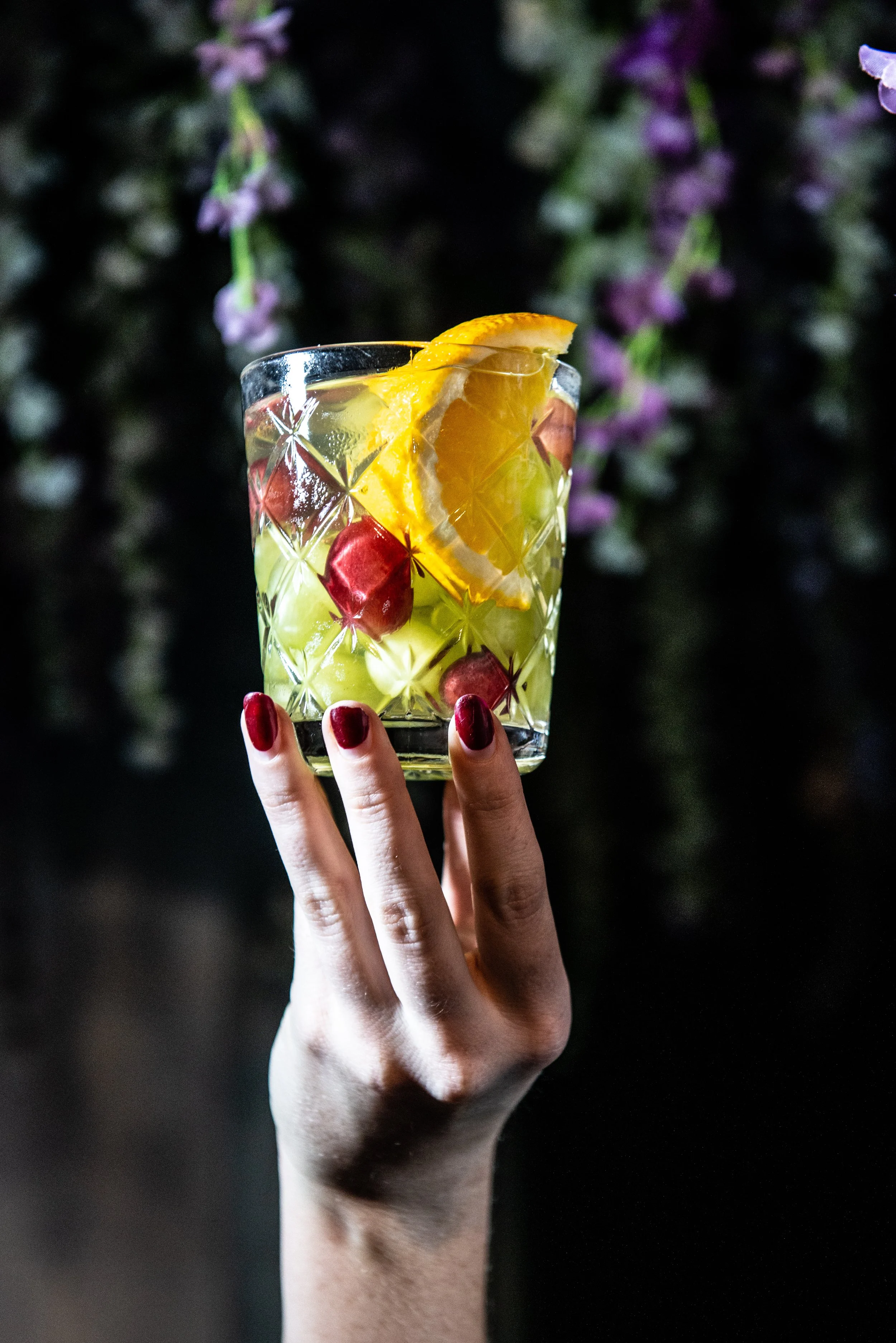 A person holding a glass of cocktail with ice, orange slices, and cherries against a dark background with blurred purple flowers.