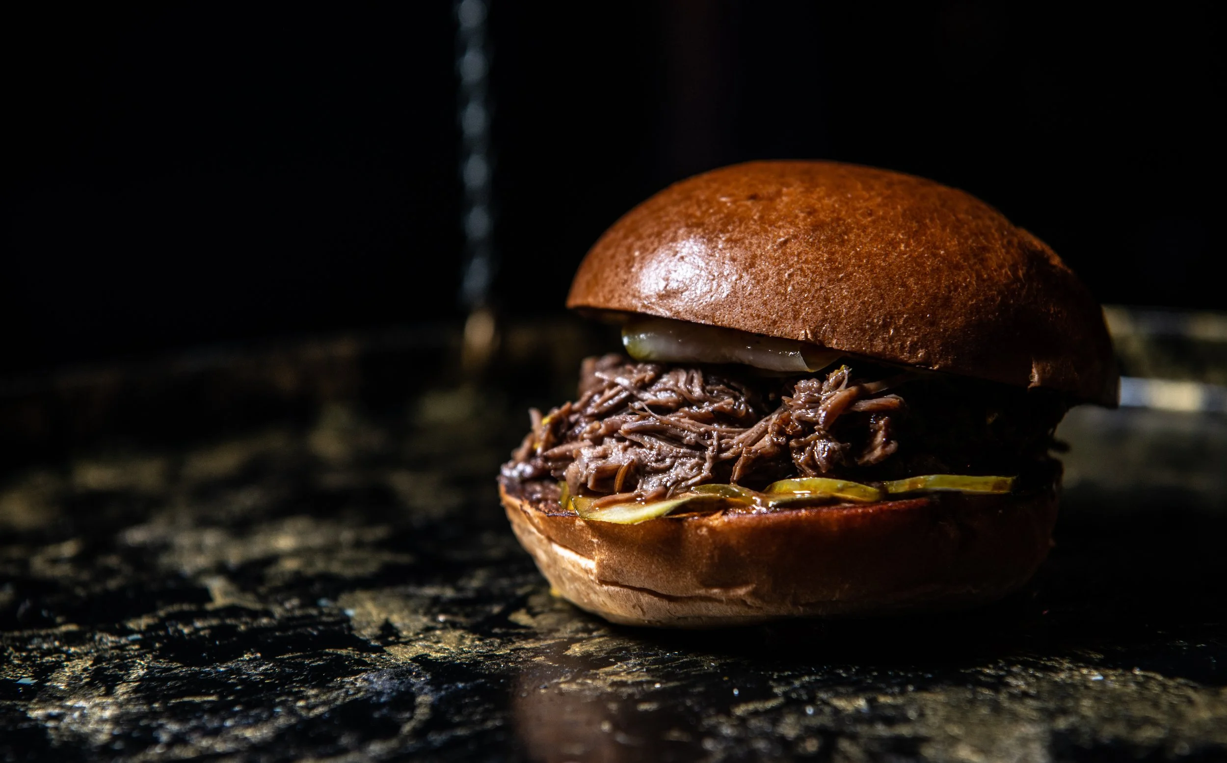 Close-up of a barbecue beef slider with shredded meat, pickles, and a brioche bun on a dark textured surface.