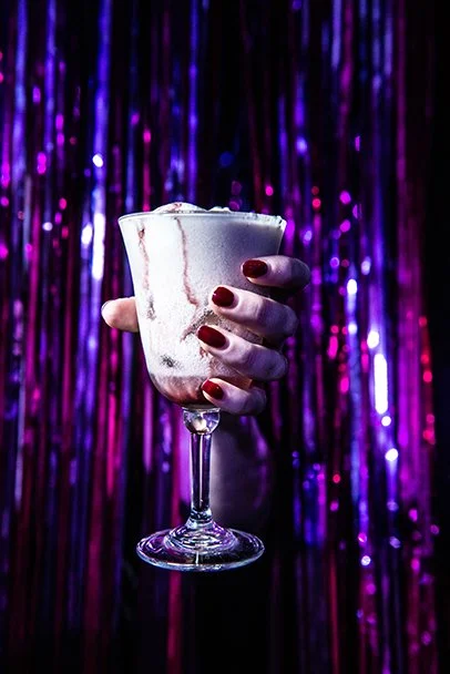 A hand with red nail polish holding a tall glass of a frothy pink cocktail in front of a purple and pink tinsel background.