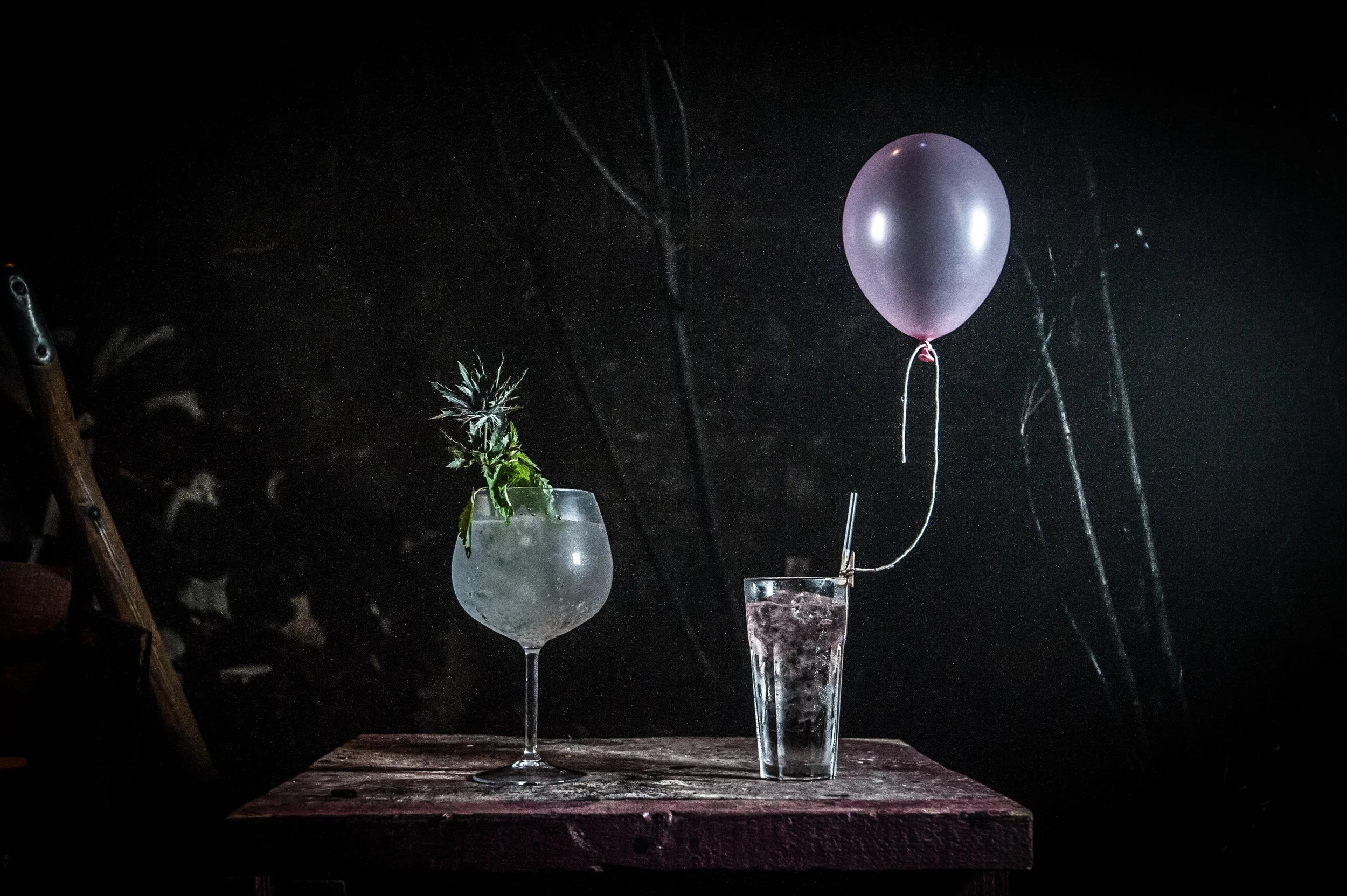 Two fairy tale champagne glasses with plants inside, one with a purple balloon behind them, placed on a wooden surface against a dark background.