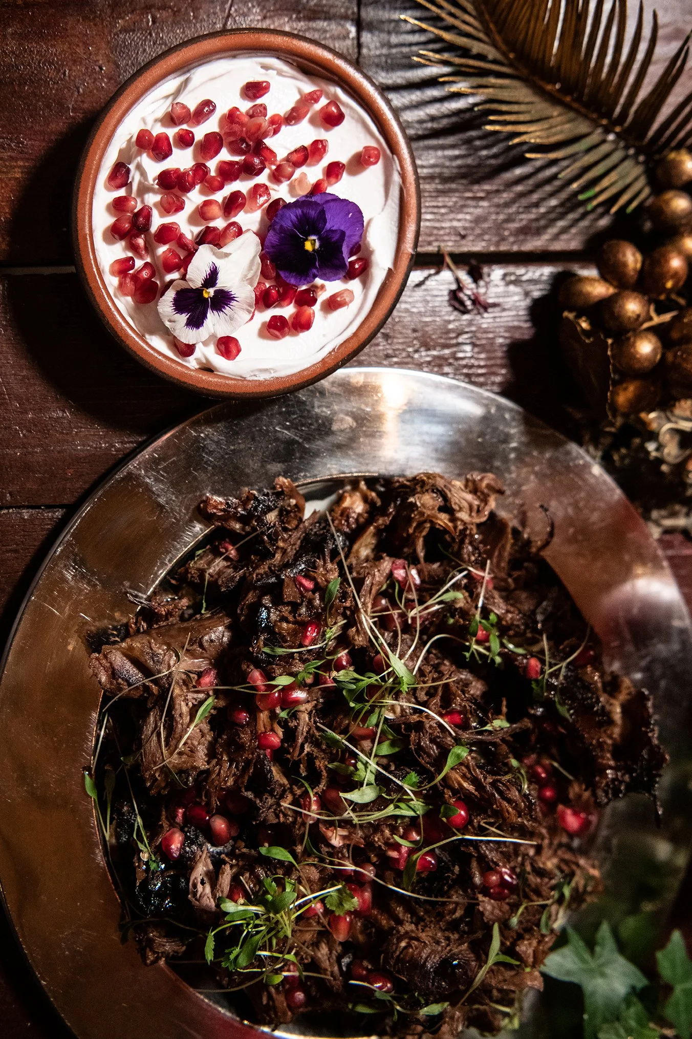 A bowl of whipped cream topped with pomegranate seeds and edible flowers, placed on a wooden surface next to a metal platter of cooked meat garnished with pomegranate seeds and herbs, with a large dried palm leaf in the background.