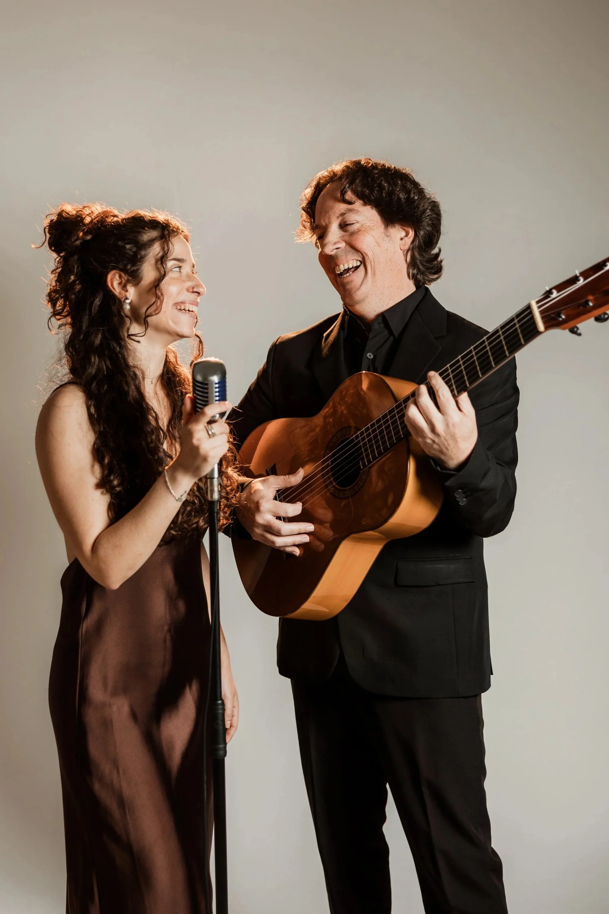 A man playing guitar and a woman singing into a microphone, both smiling and looking at each other.