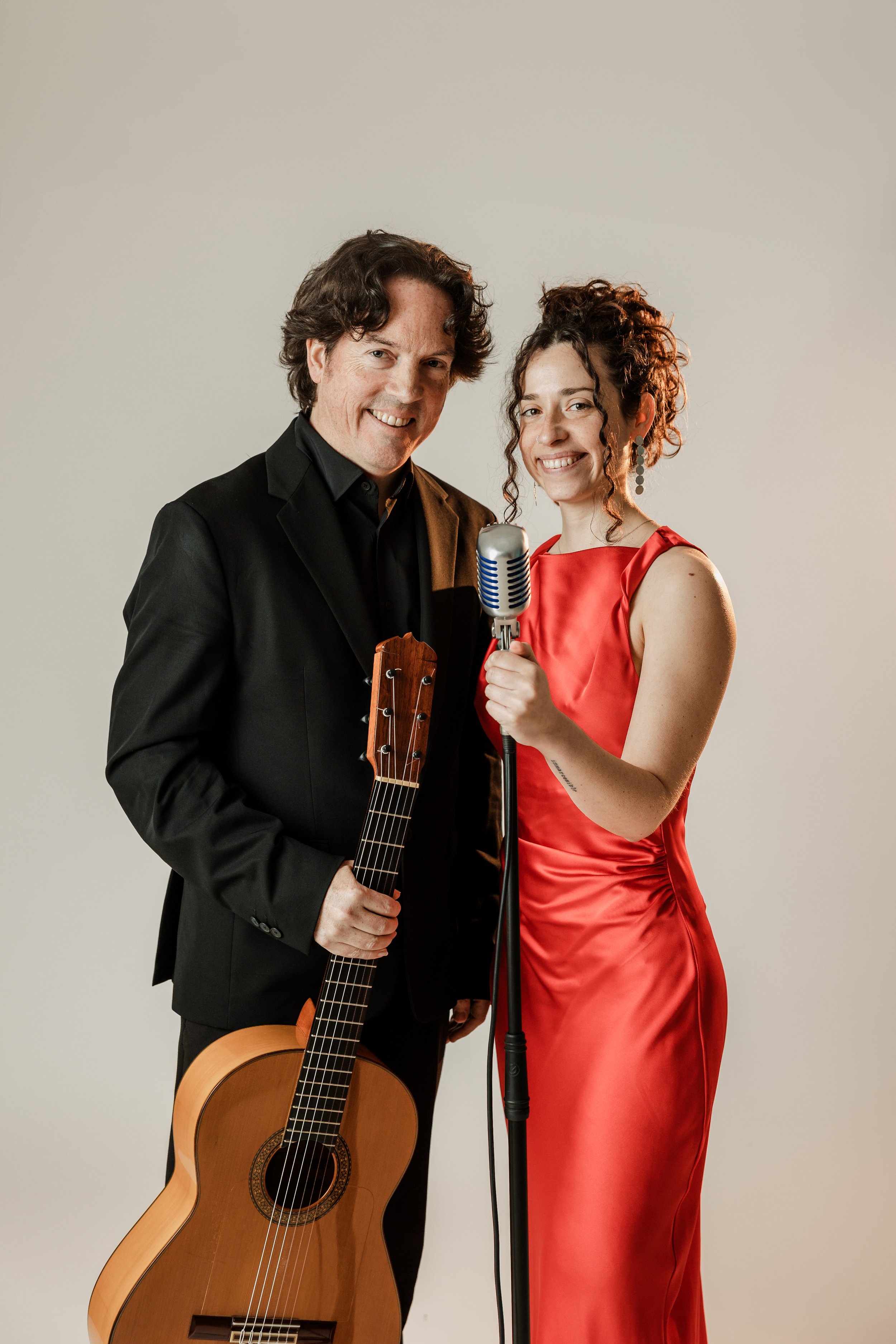 A man and a woman standing together, the man holding a flamenco guitar and the woman holding a microphone, both smiling at the camera, against a plain light background.