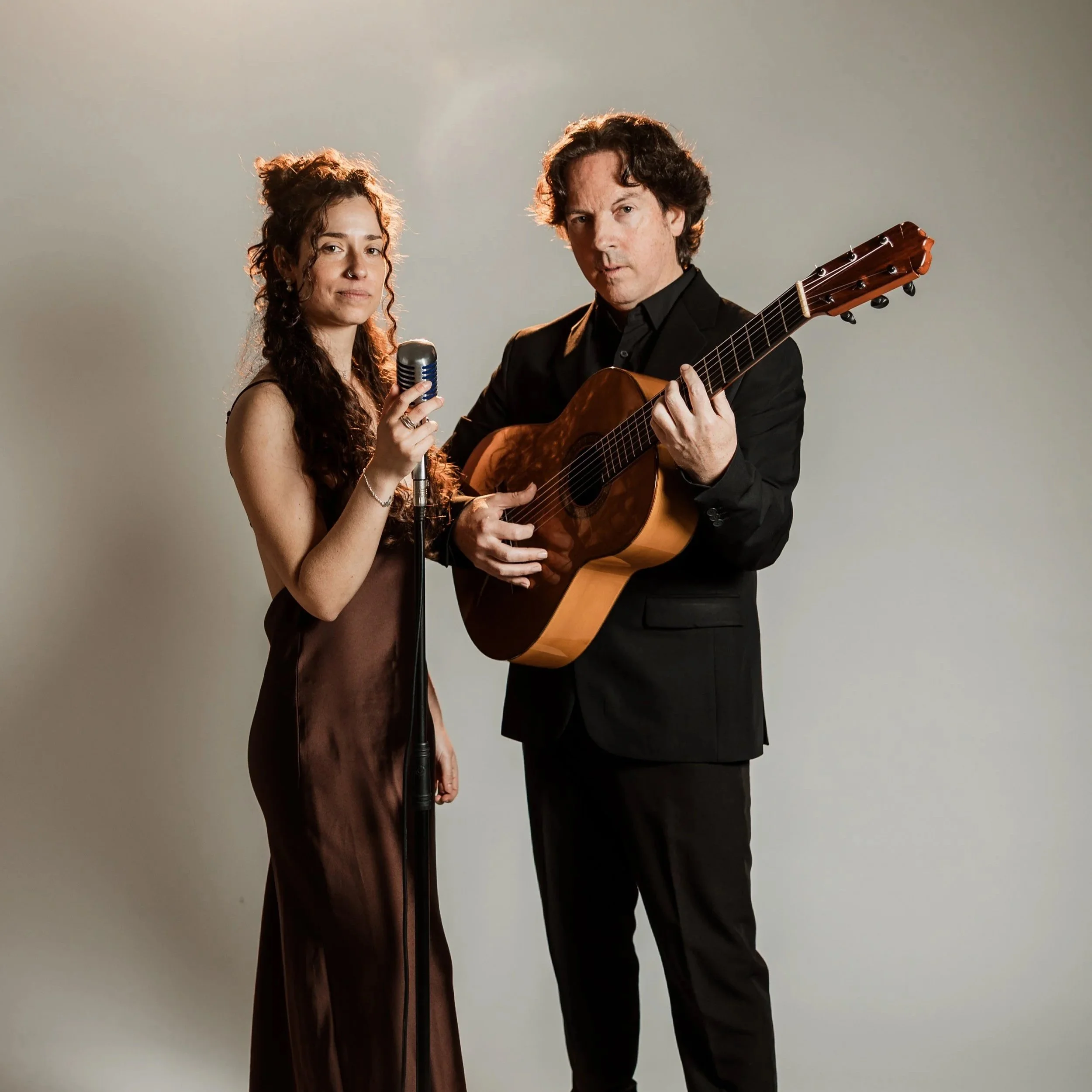A woman holding a microphone standing next to a man playing an acoustic guitar, both dressed in formal attire, in front of a plain background.