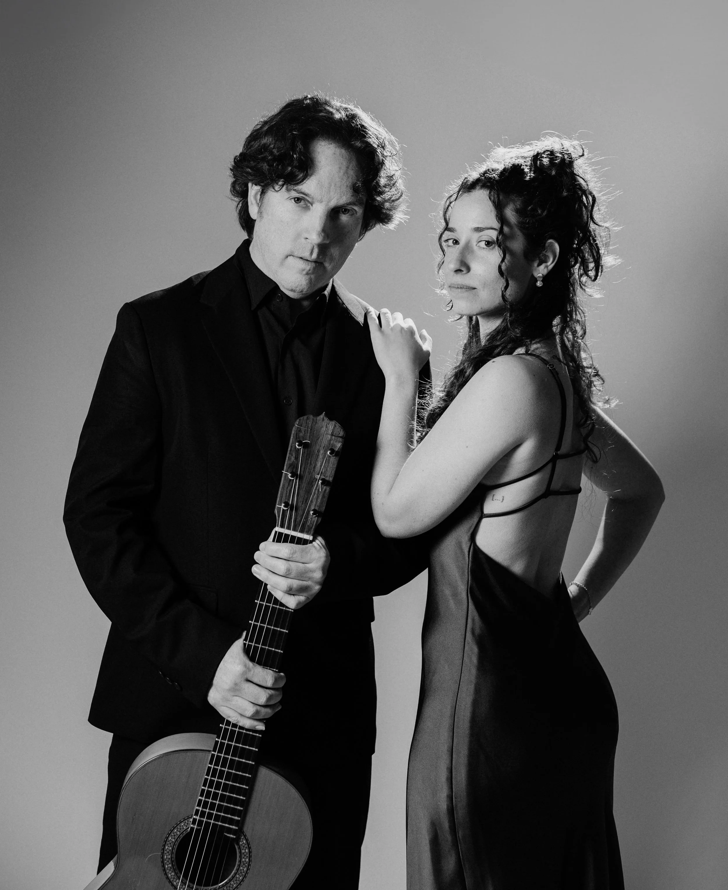 Black and white photo of a man in a suit holding a Spanish guitar and a woman in a satin dress standing close together with the woman