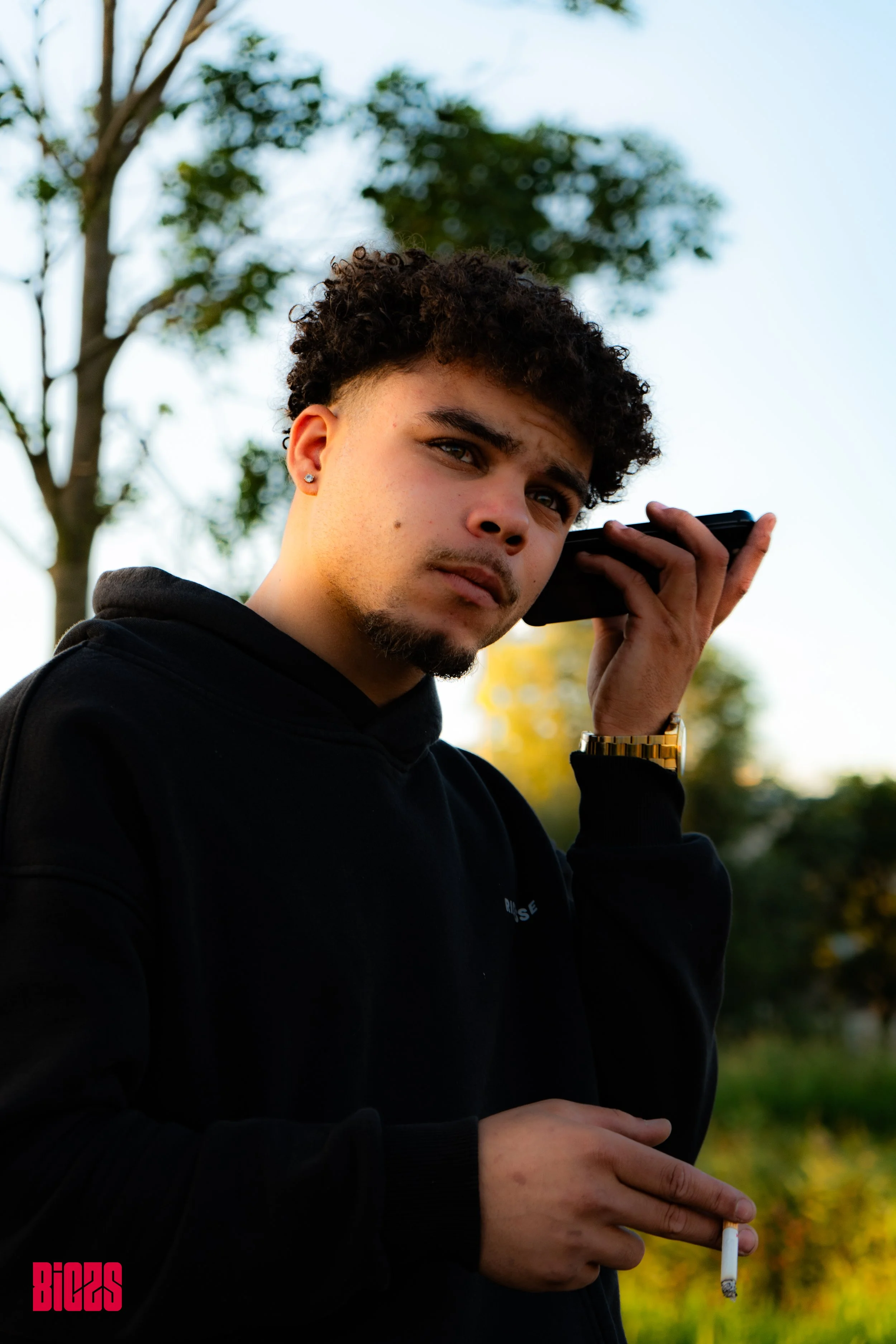 A young man with curly hair and a goatee holding a cellphone to his ear and a cigarette in his hand, standing outdoors with trees and a clear sky in the background.