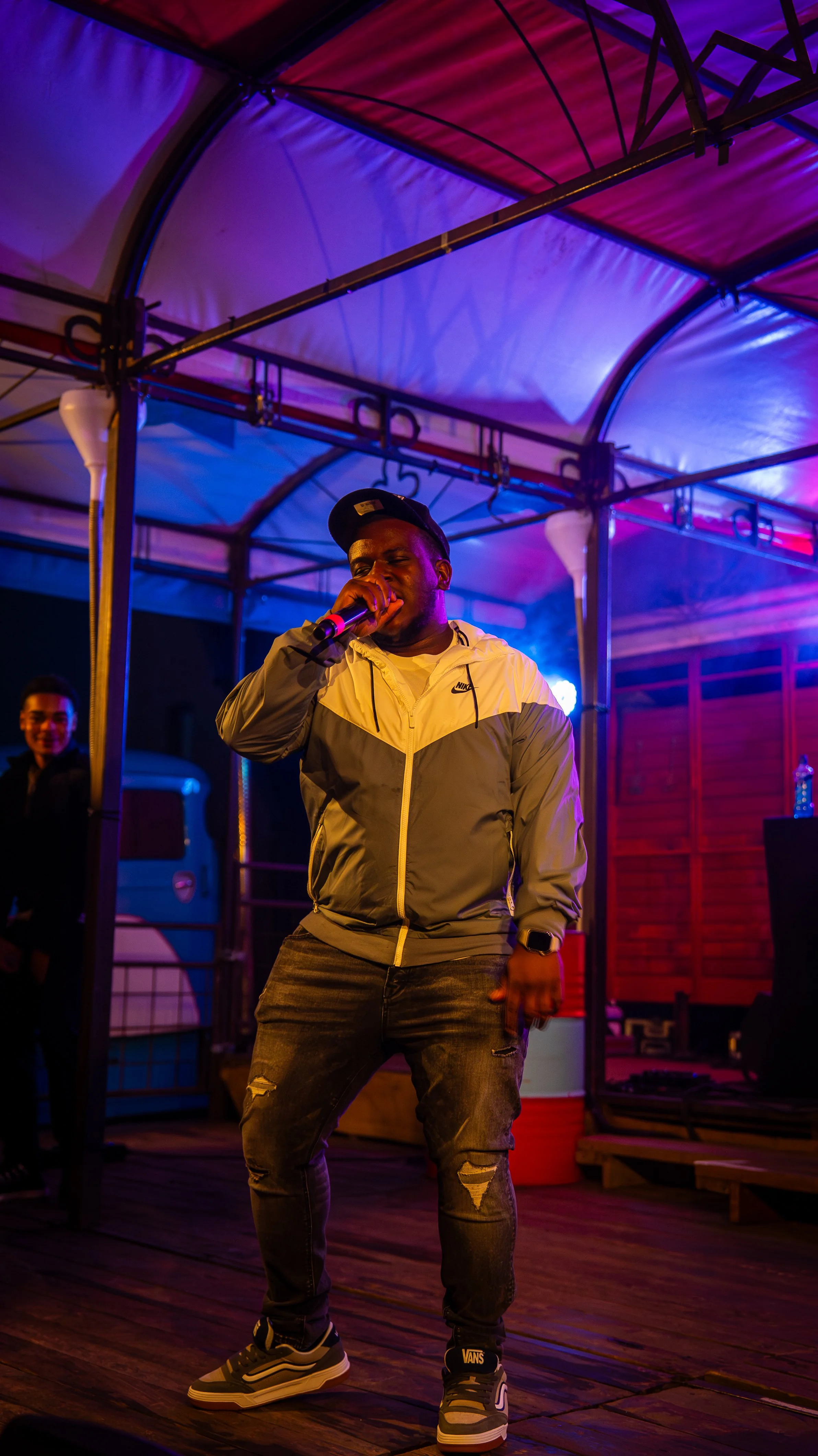 A male performer singing into a microphone on a small stage at night, under colorful lighting, with another person smiling in the background.