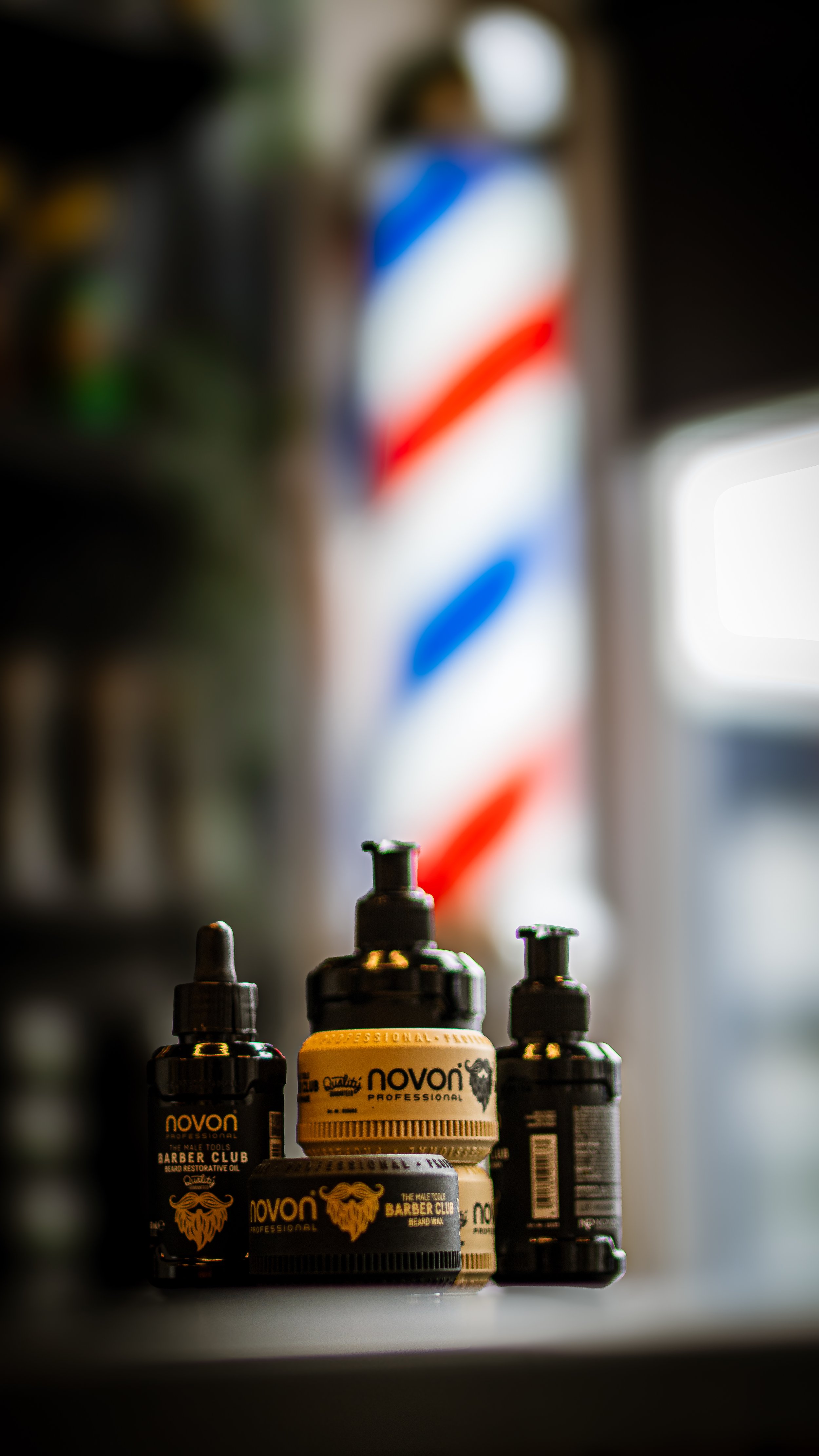 A collection of Novon beard grooming products, including oil, wax, and balm, in front of a blurred barber pole with red, white, and blue stripes.