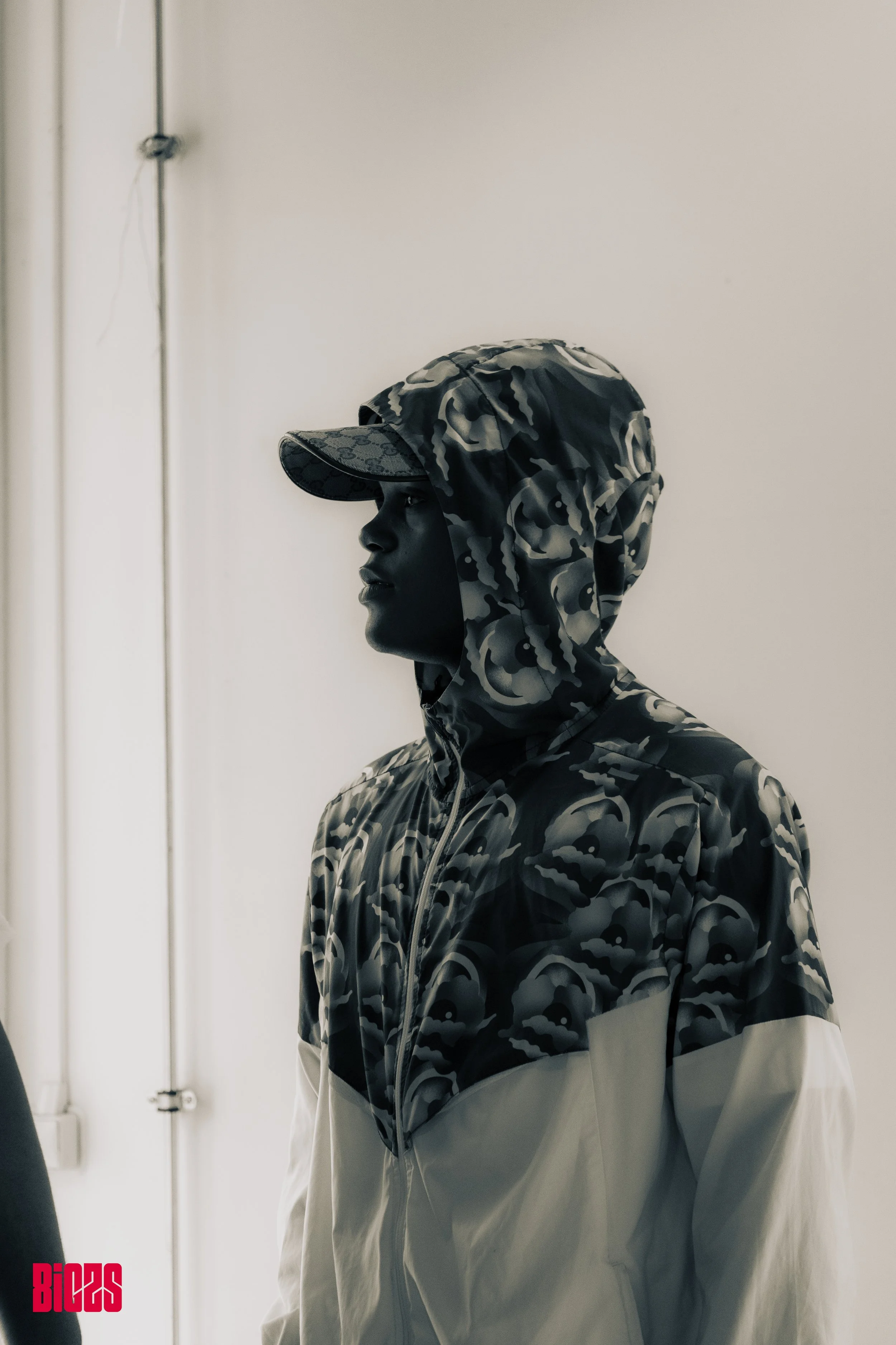 Side view of a person wearing a camo hooded jacket and cap, standing indoors near a wall.