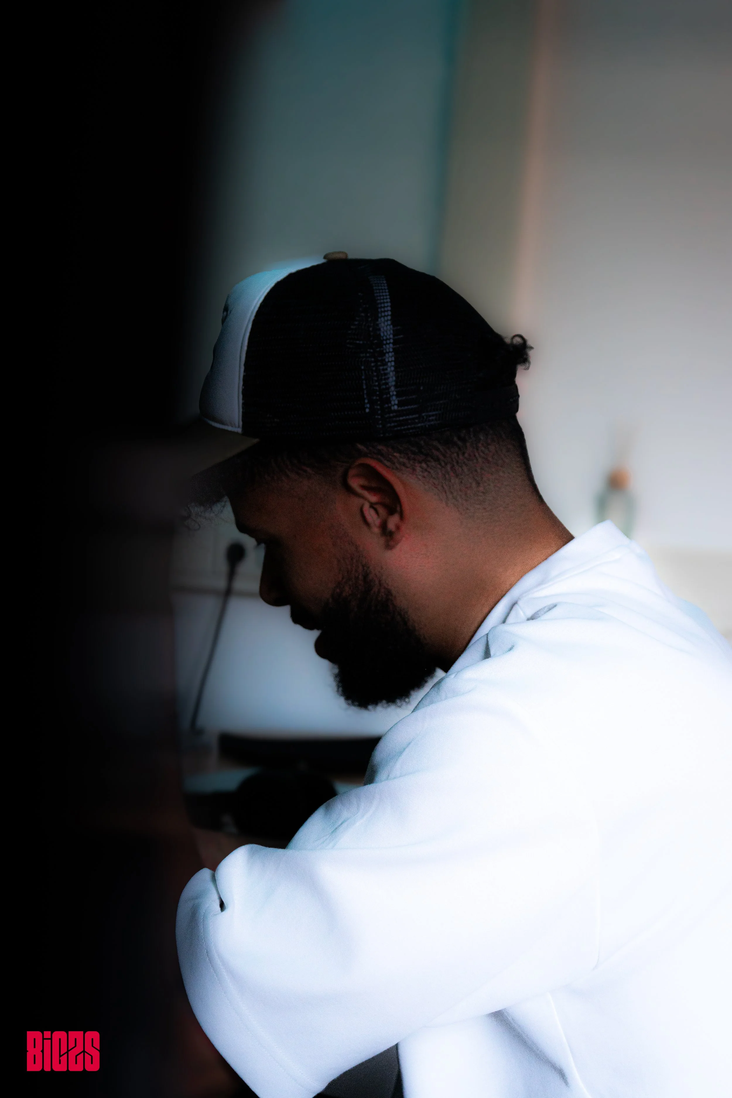 A man with a beard and a black and white trucker hat sitting in a dimly lit room, looking down, wearing a white shirt.