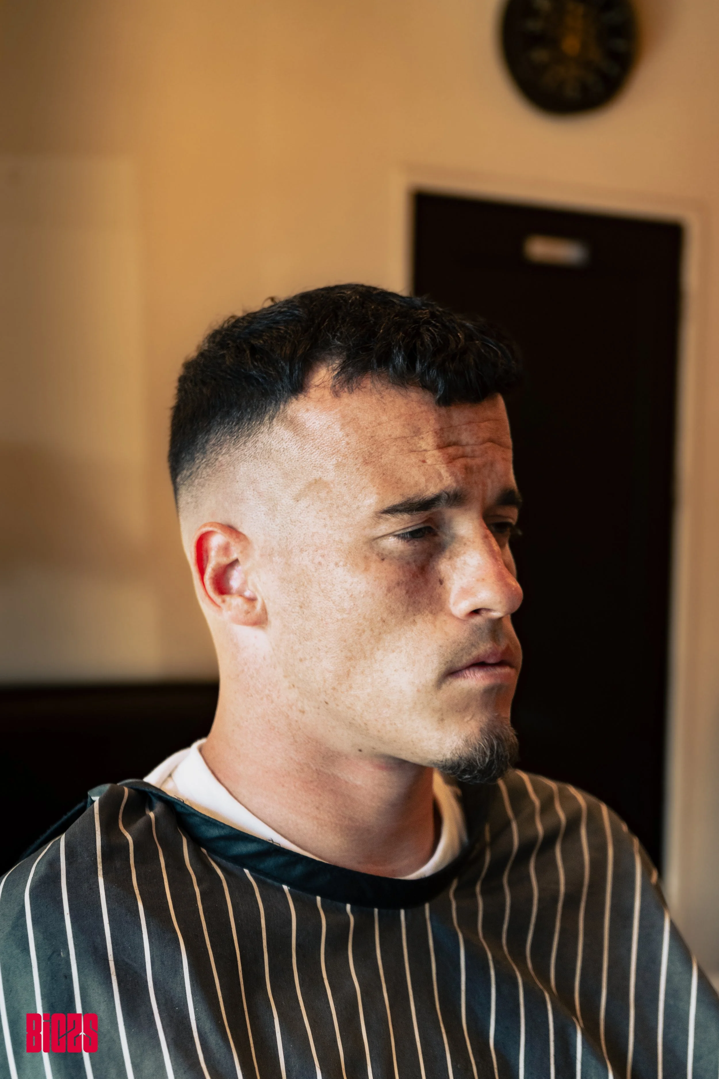 A young man with a short fade haircut sitting in a barbershop chair, looking down with a serious expression, wearing a black and white pinstripe barber cape. The background features a beige wall, a clock, and a partly open door.