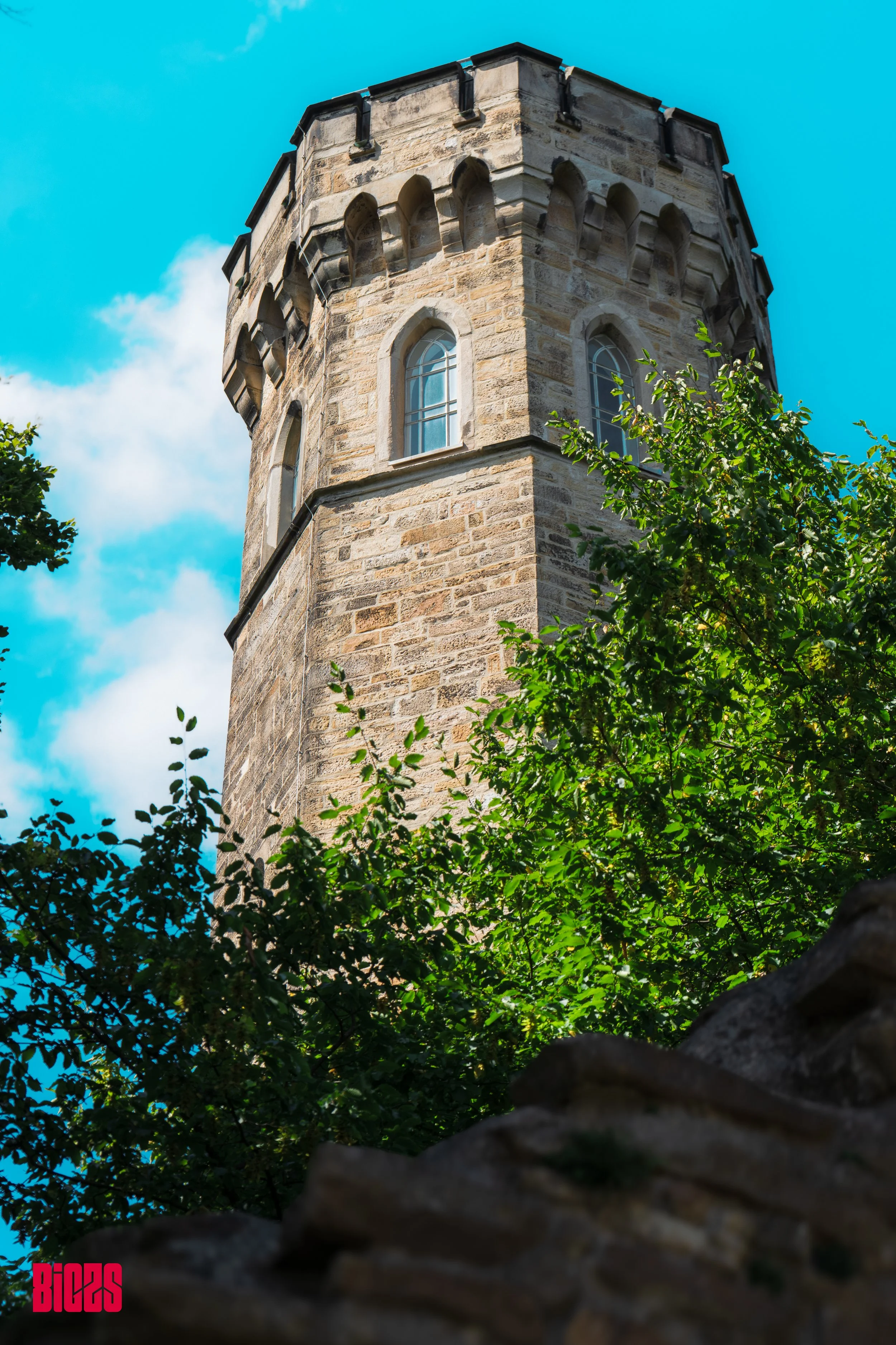A tall stone tower with narrow arched windows, surrounded by green trees against a blue sky with clouds.