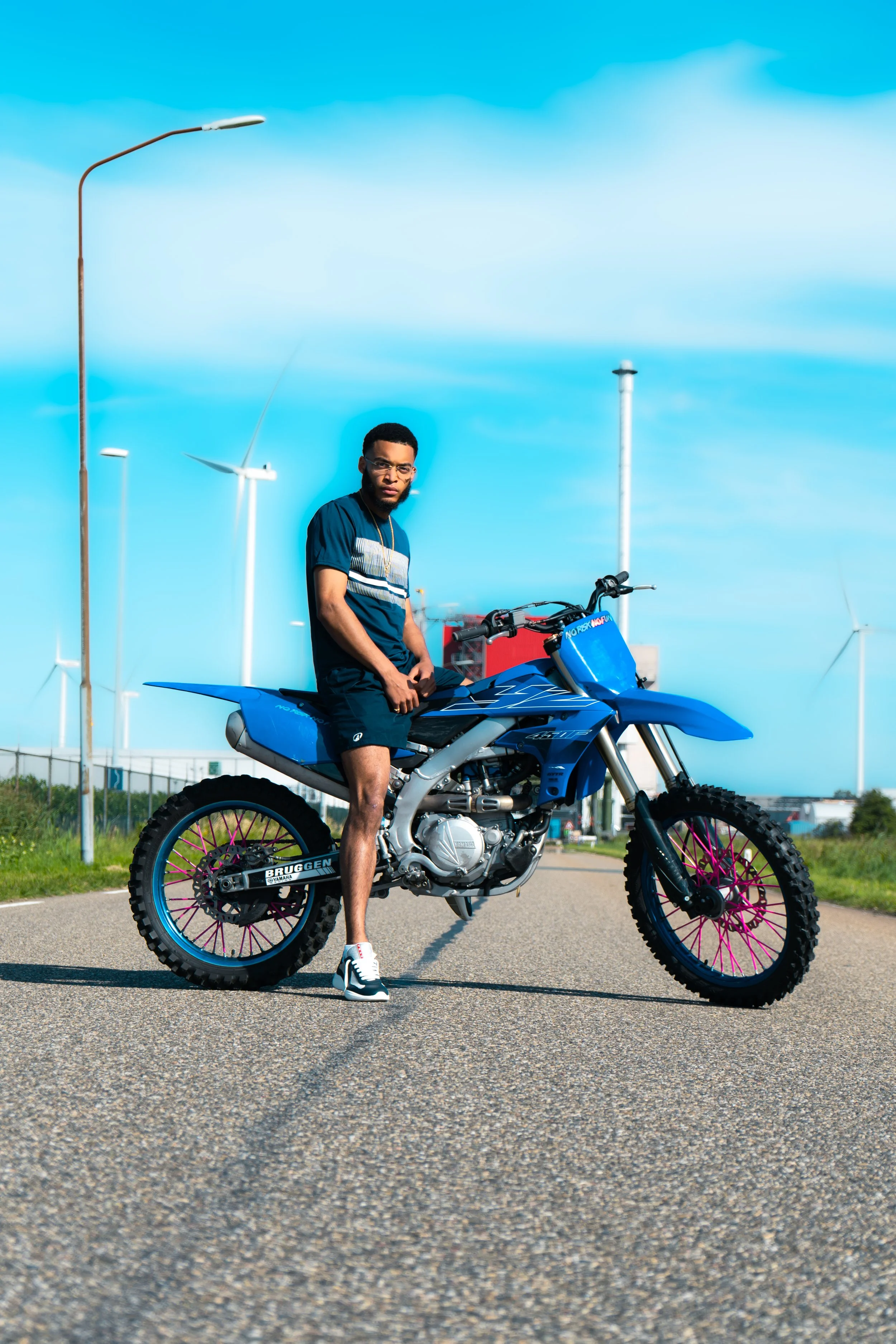 A young man standing next to a blue off-road motorcycle on a paved road, with wind turbines and a blue sky in the background.