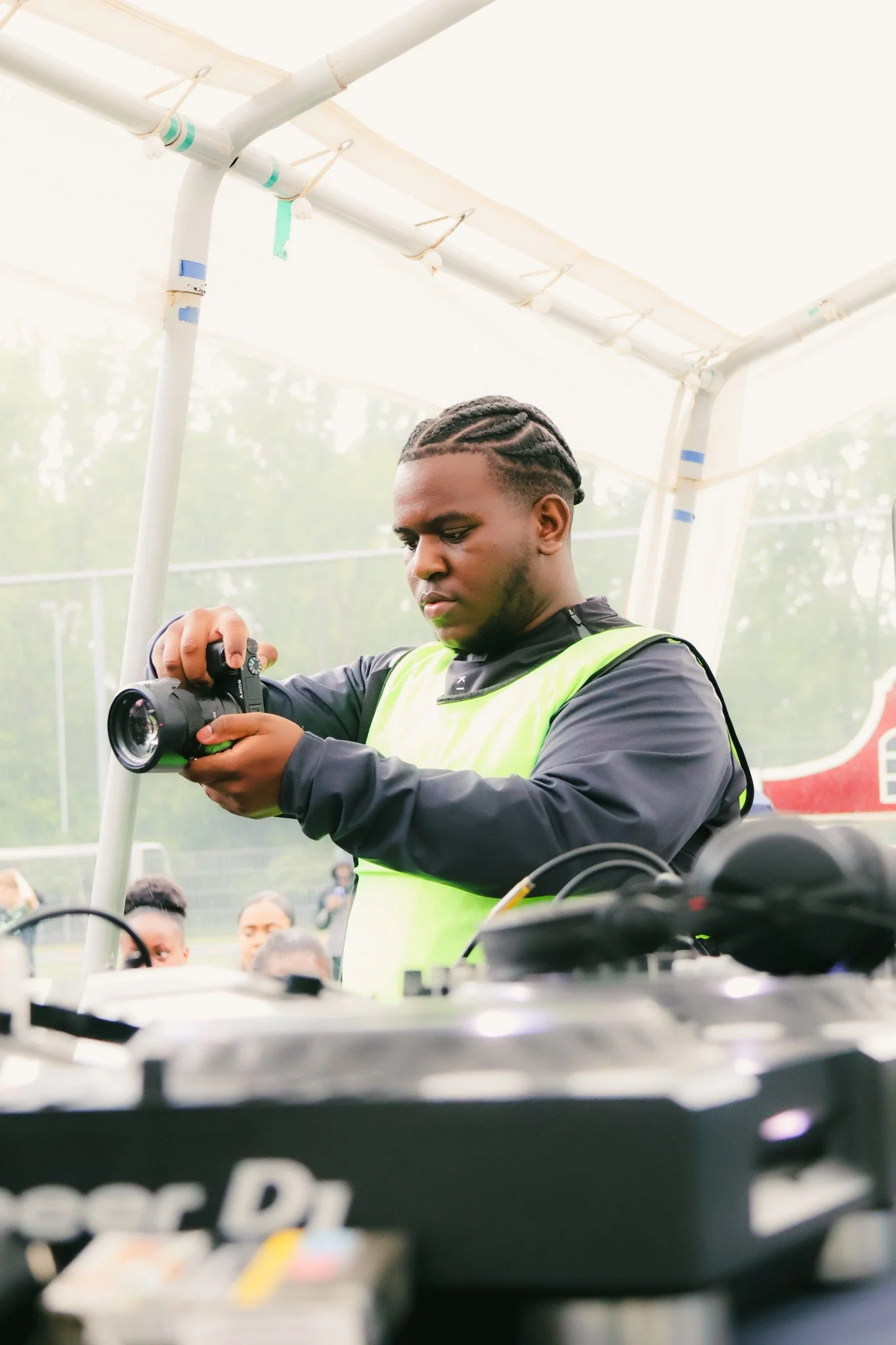 A young man wearing a neon green safety vest and black long sleeve shirt is looking at a camera. He is standing under a white tent with a metal frame. There are people in the background and equipment on the table in front of him.