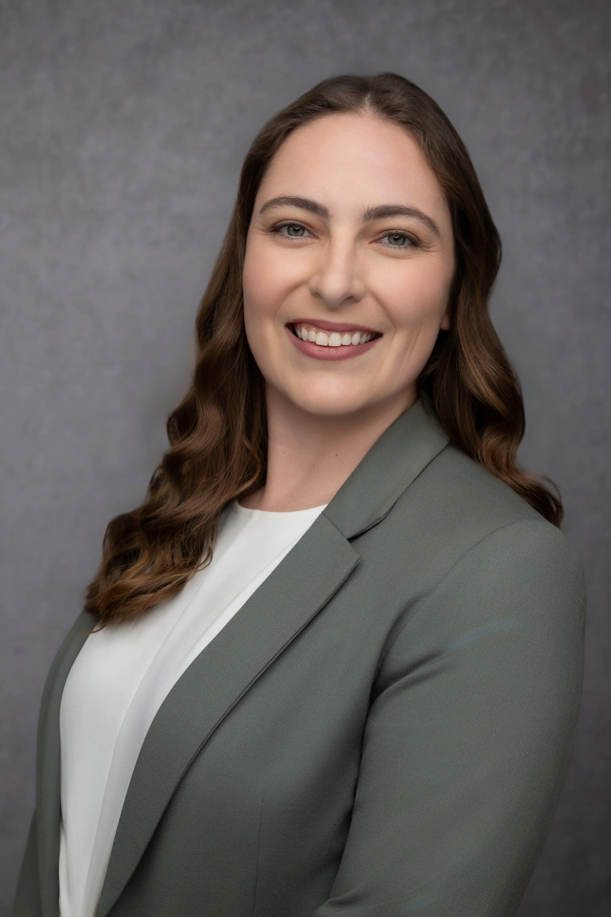 A woman with long wavy brown hair smiling in a professional headshot, wearing a gray blazer over a white blouse against a gray background.