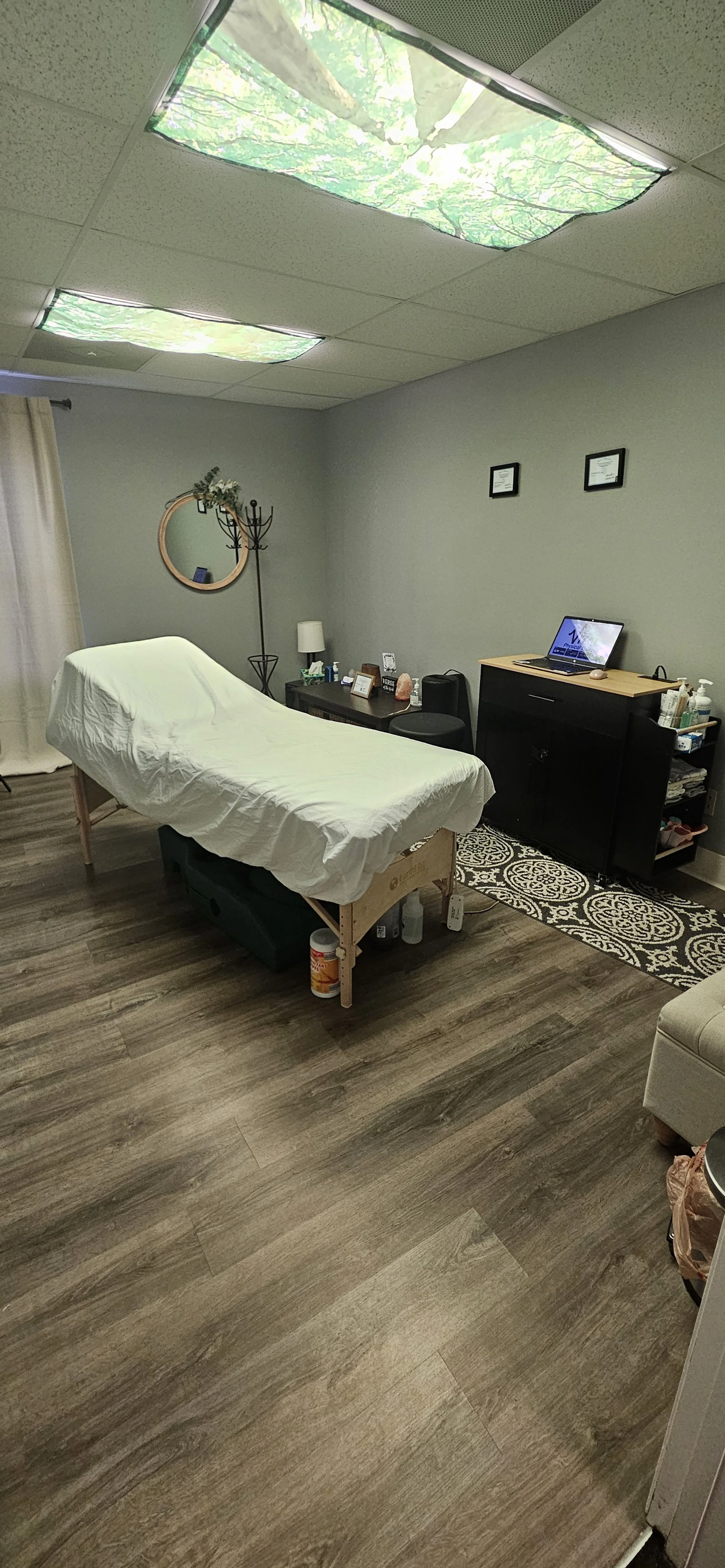 A treatment room in St. Charles, MO with a massage table covered in a white sheet, a black cabinet with tools and supplies, a small rug, a window looking to outdoors, and ceiling lights that have a forest scene projected on them.