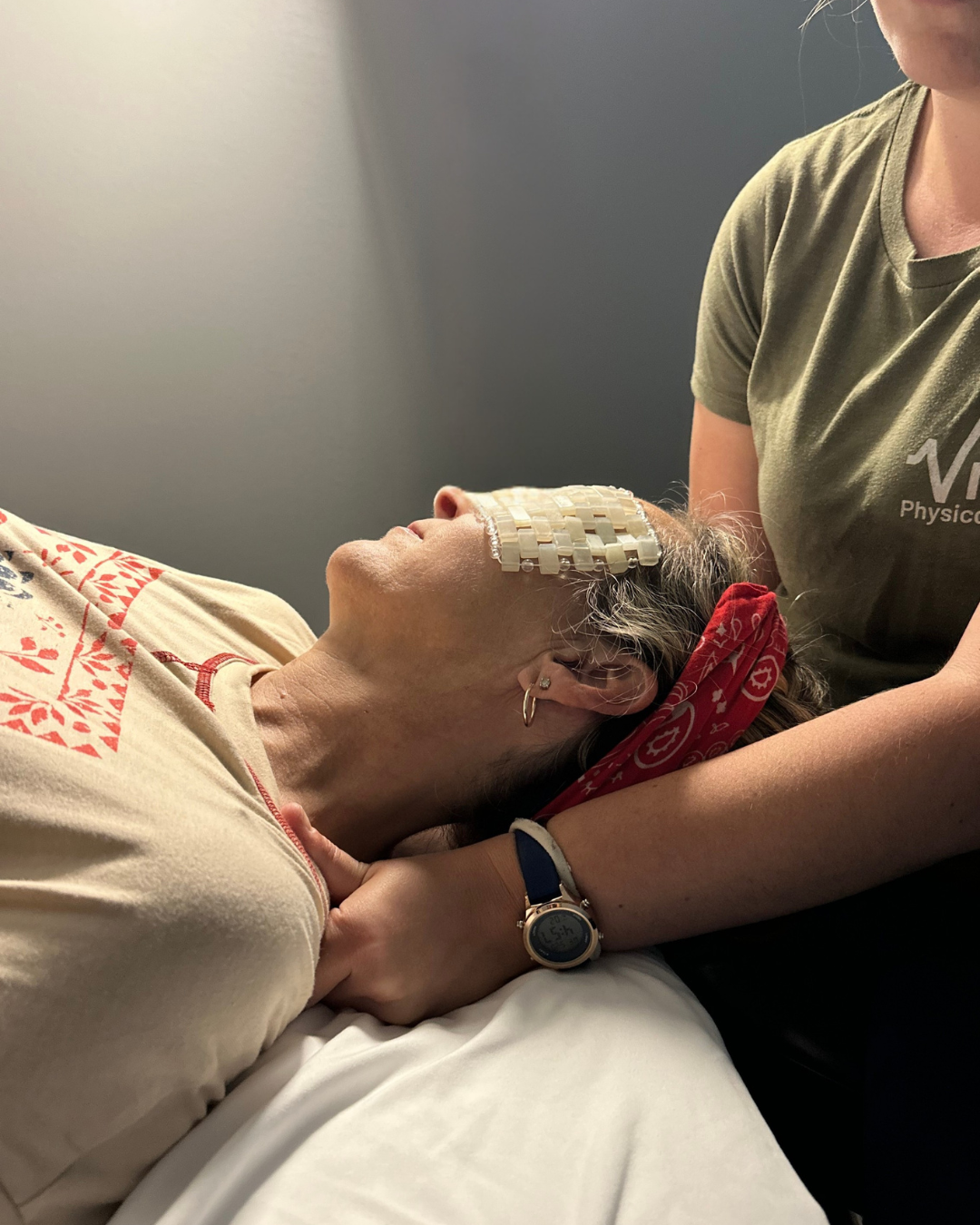 A woman lying in a hospital bed with a headband and an eye patch connected to a device on her forehead, being supported by another person.