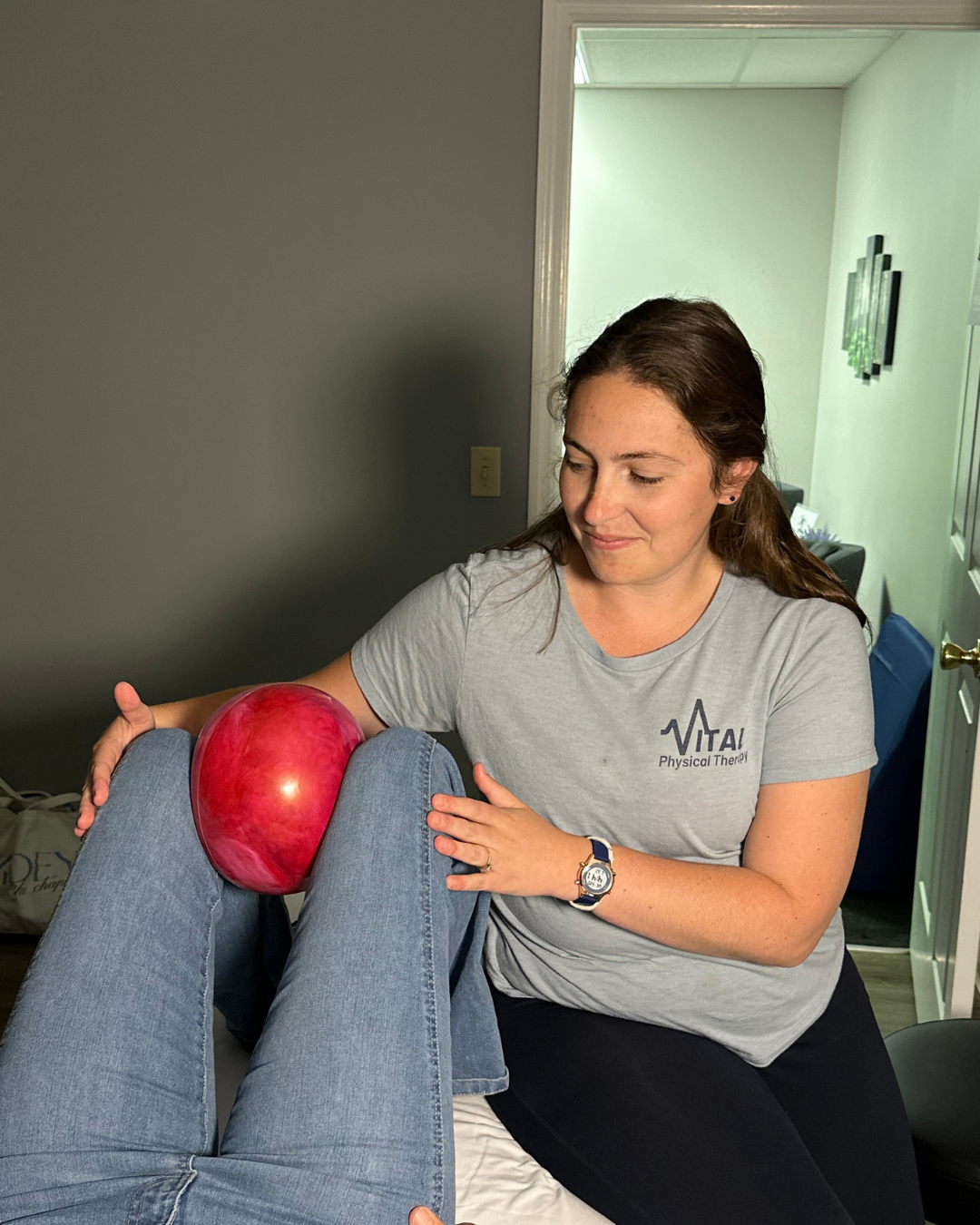 A physical therapist holds a red exercise ball against a patient's knee during therapy in a treatment room.