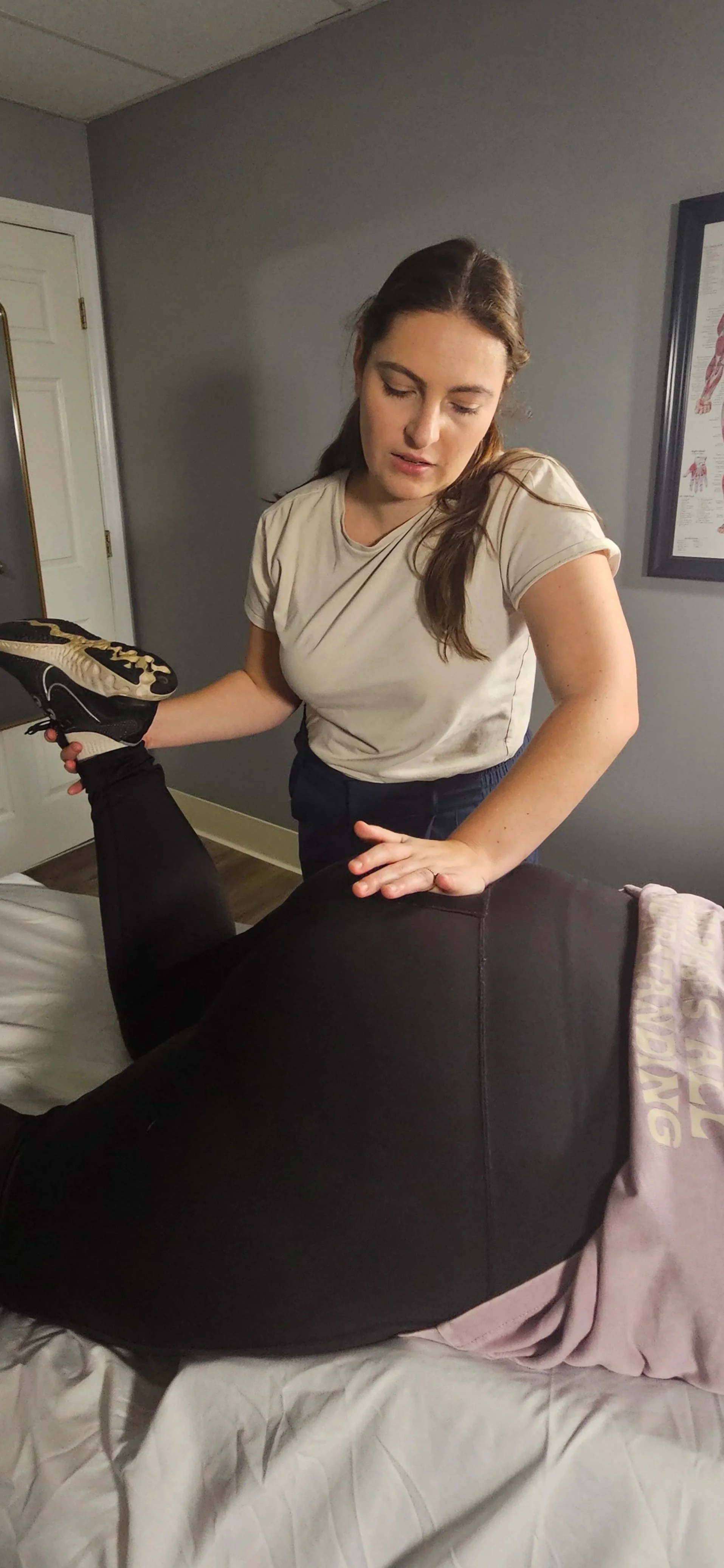 A woman in a beige t-shirt is performing a physical therapy exercise on a person lying face down on a massage table, lifting their leg with her hand supporting their thigh.