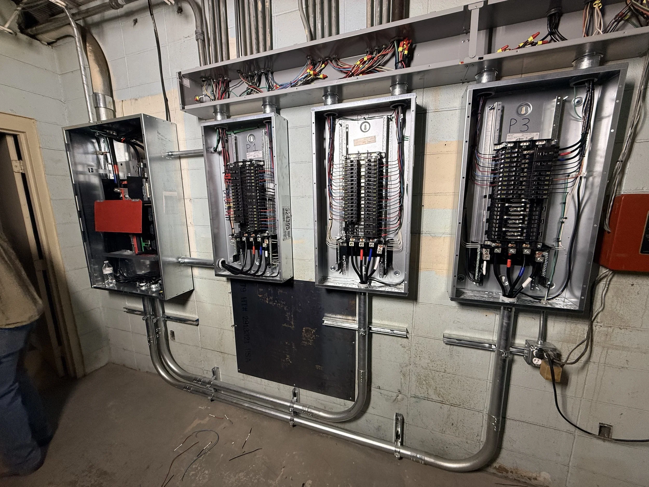 Electrical panels with wiring and circuit breakers mounted on a brick wall in an industrial setting.