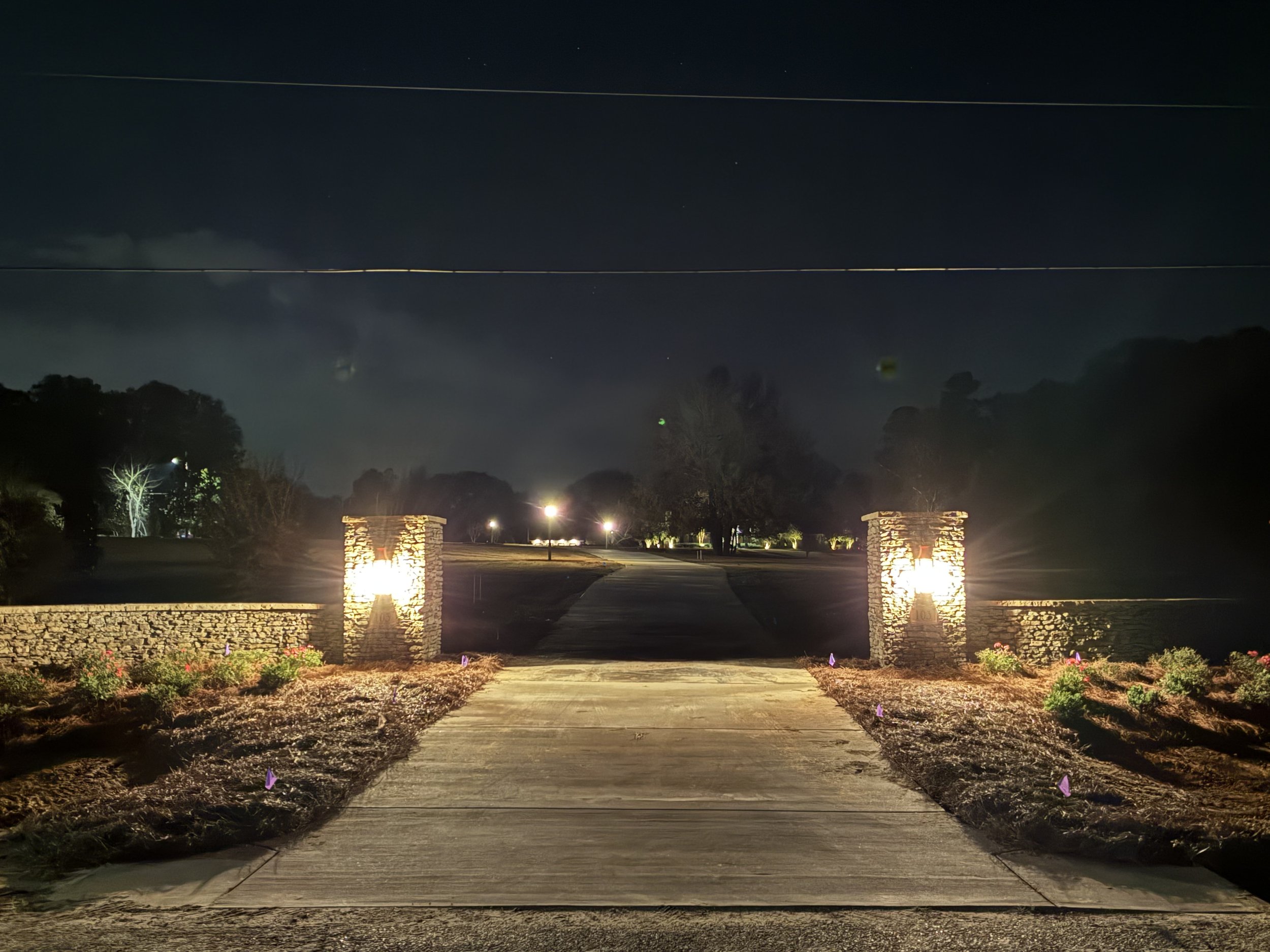 Nighttime view of a driveway leading to a park or garden, with stone pillar entrance, lit with outdoor lights, trees and open space in the background.