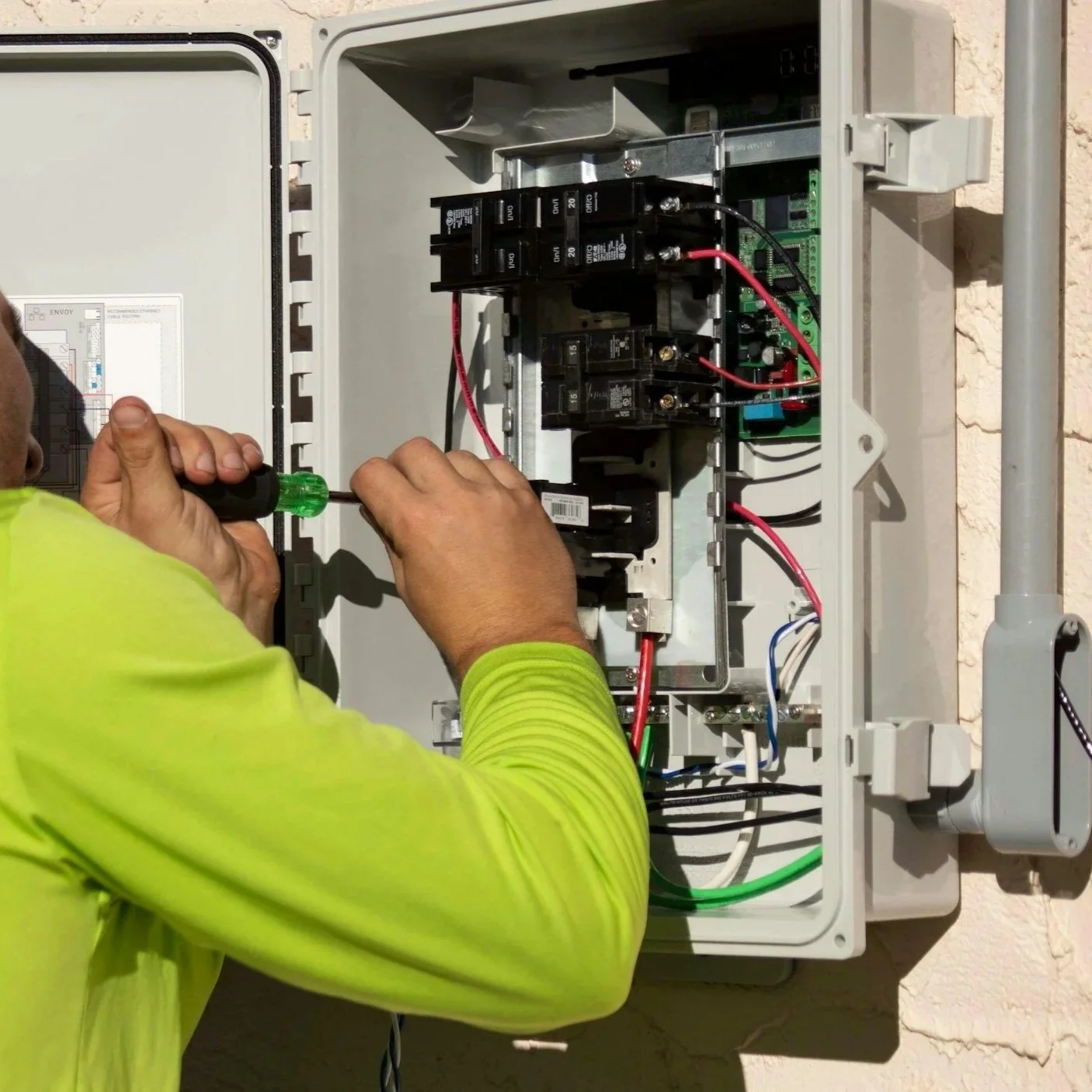 A technician wearing a bright yellow shirt working on an outdoor electrical panel with a screwdriver.