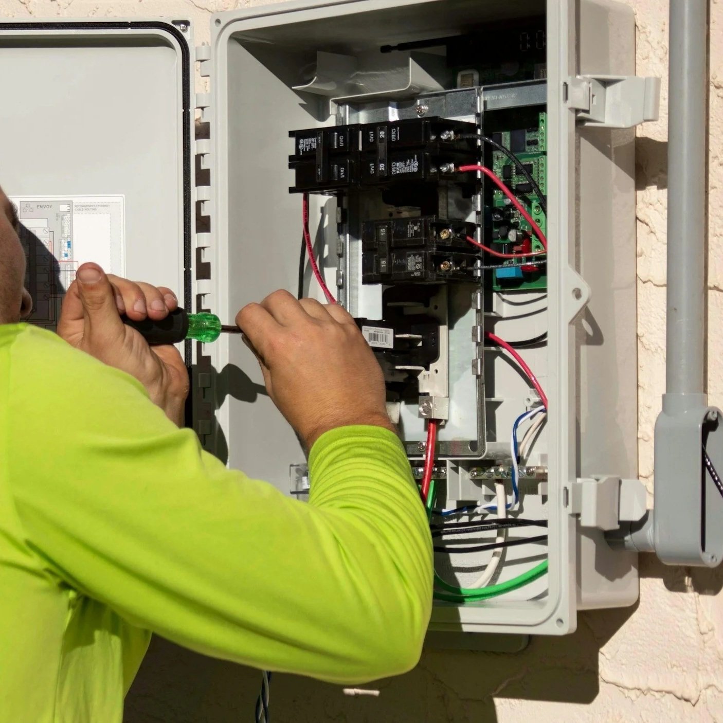 A technician working on an electrical control panel with various switches, wires, and circuit breakers inside a gray outdoor box.