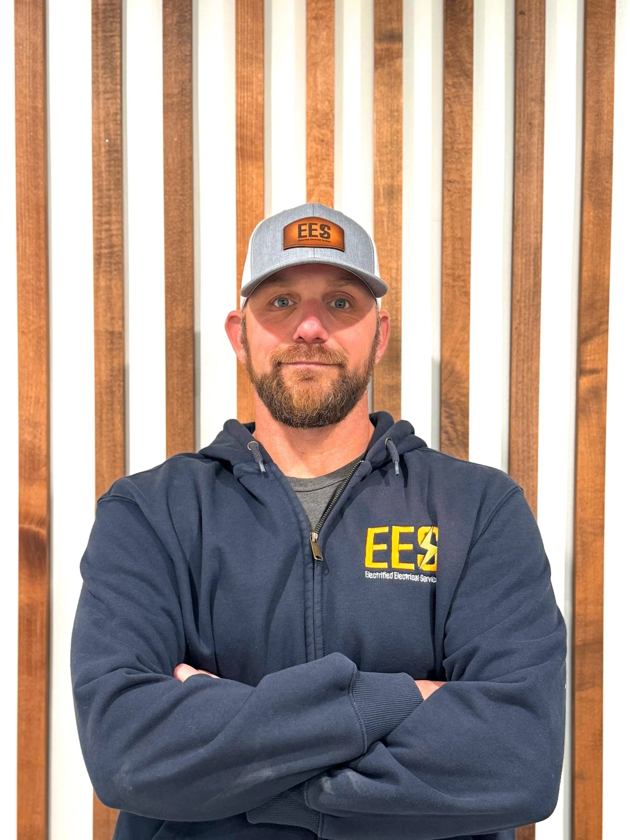 A man with a beard and mustache wearing a gray cap with EES logo and a navy blue jacket with EES logo, crossing arms in front of a wooden and white slat wall.