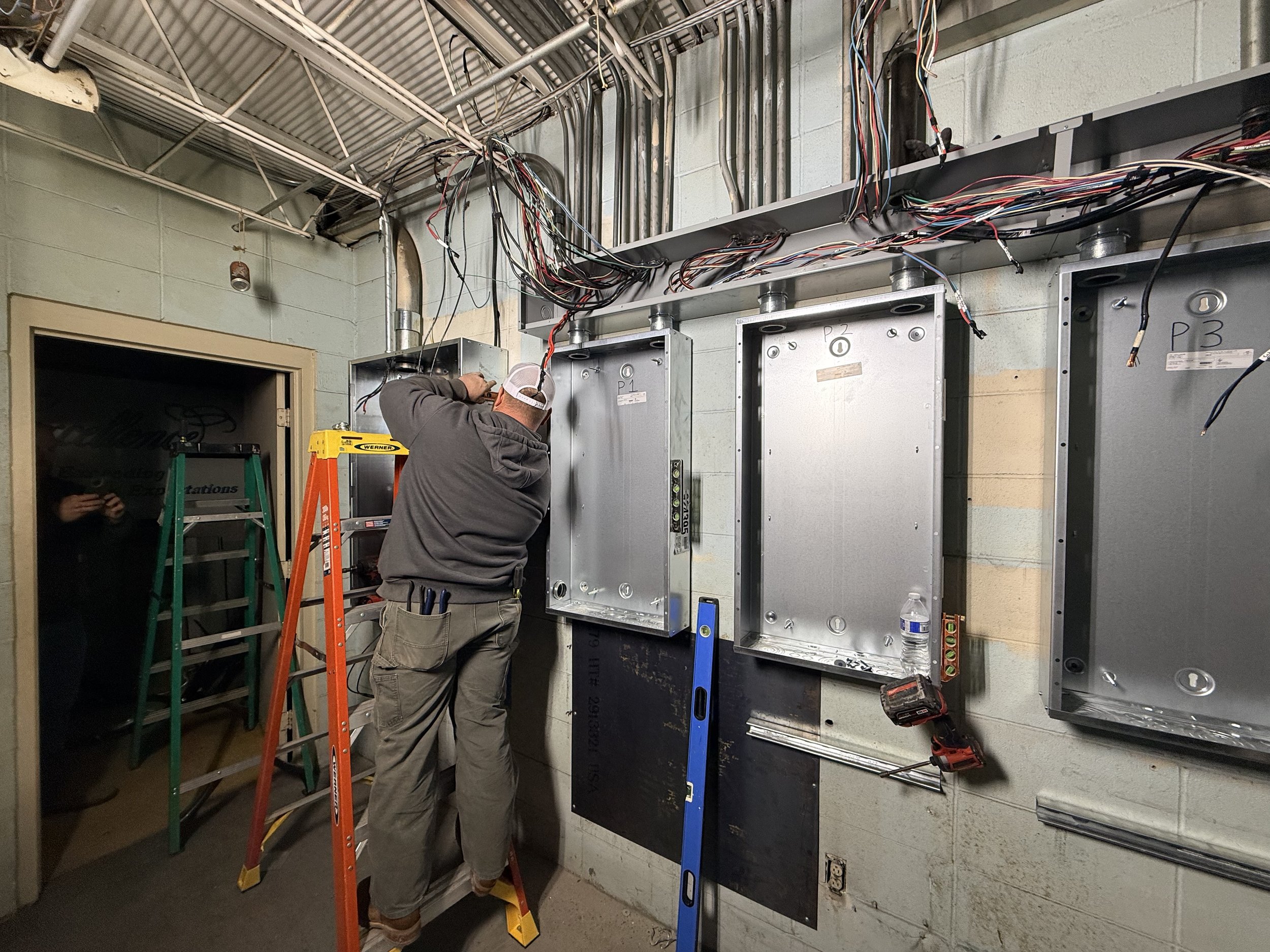 A worker standing on an orange step ladder installing or repairing electrical wiring inside an electrical box on a wall, with multiple wires hanging above, and tools such as a level and a cordless drill nearby.