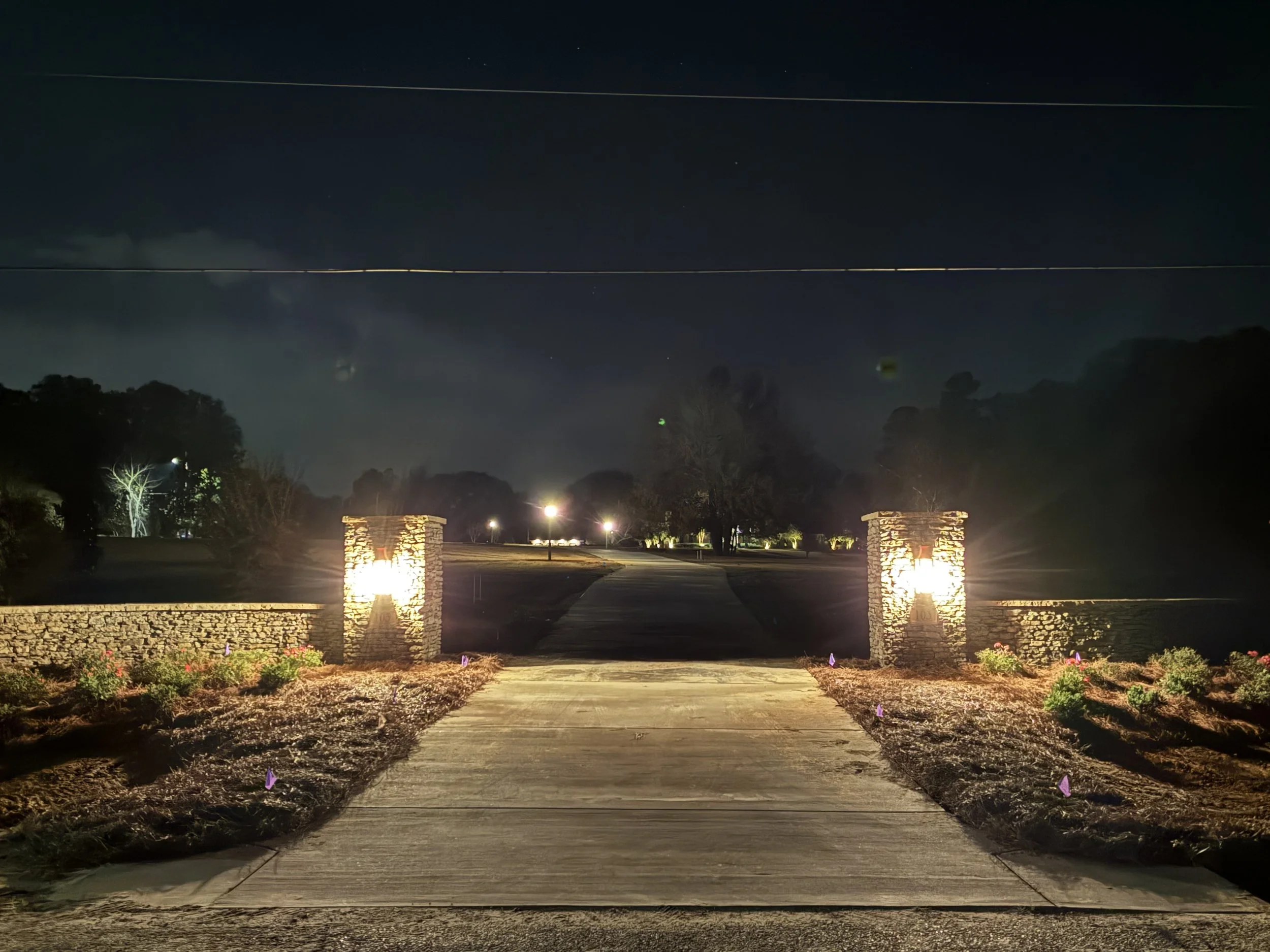 Nighttime view of a driveway entrance with stone pillars topped with lights, a sidewalk leading into a park or estate, and trees illuminated in the background under a dark sky.