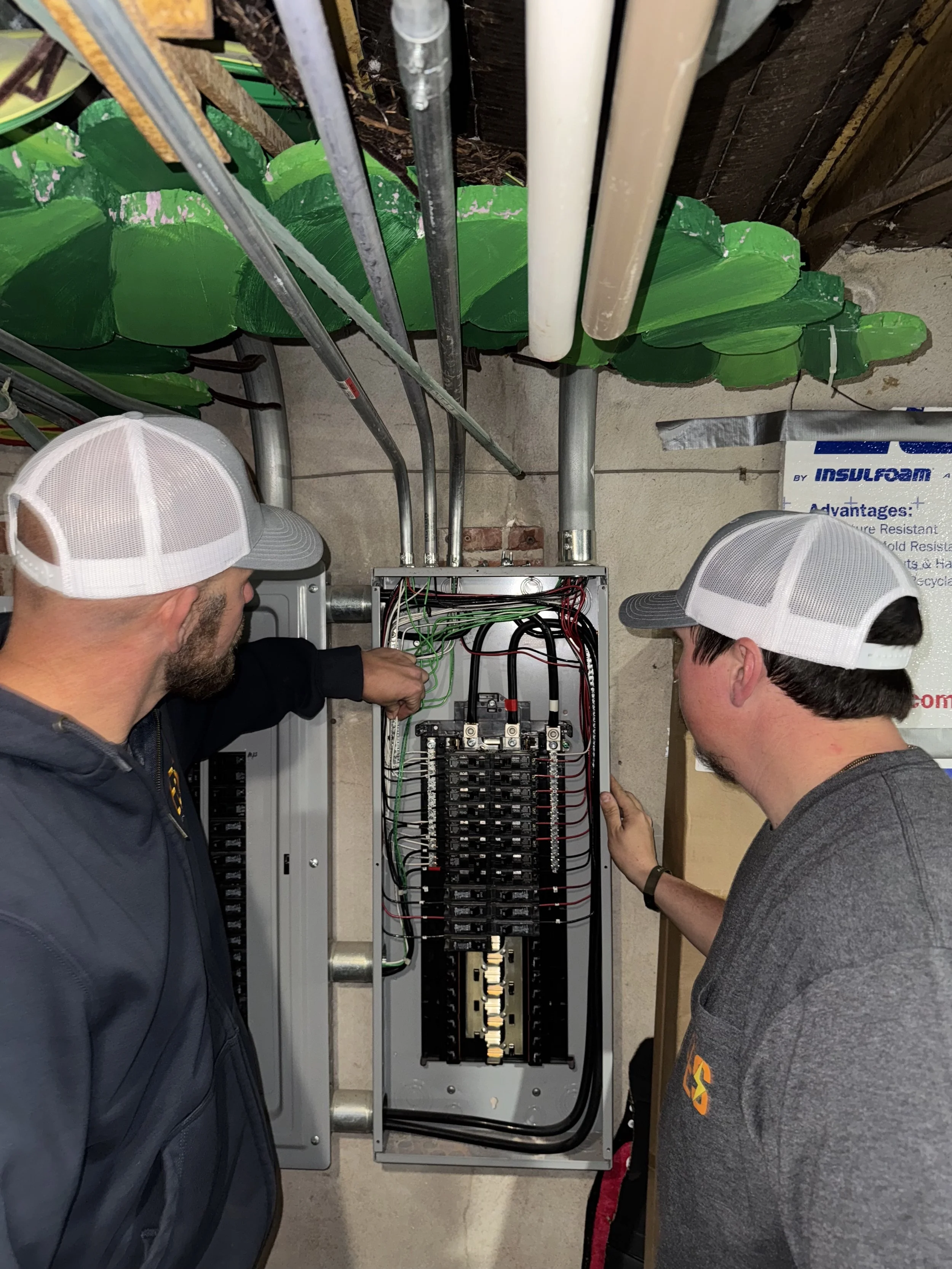 Two men in baseball caps inspecting an electrical circuit box with wires inside.