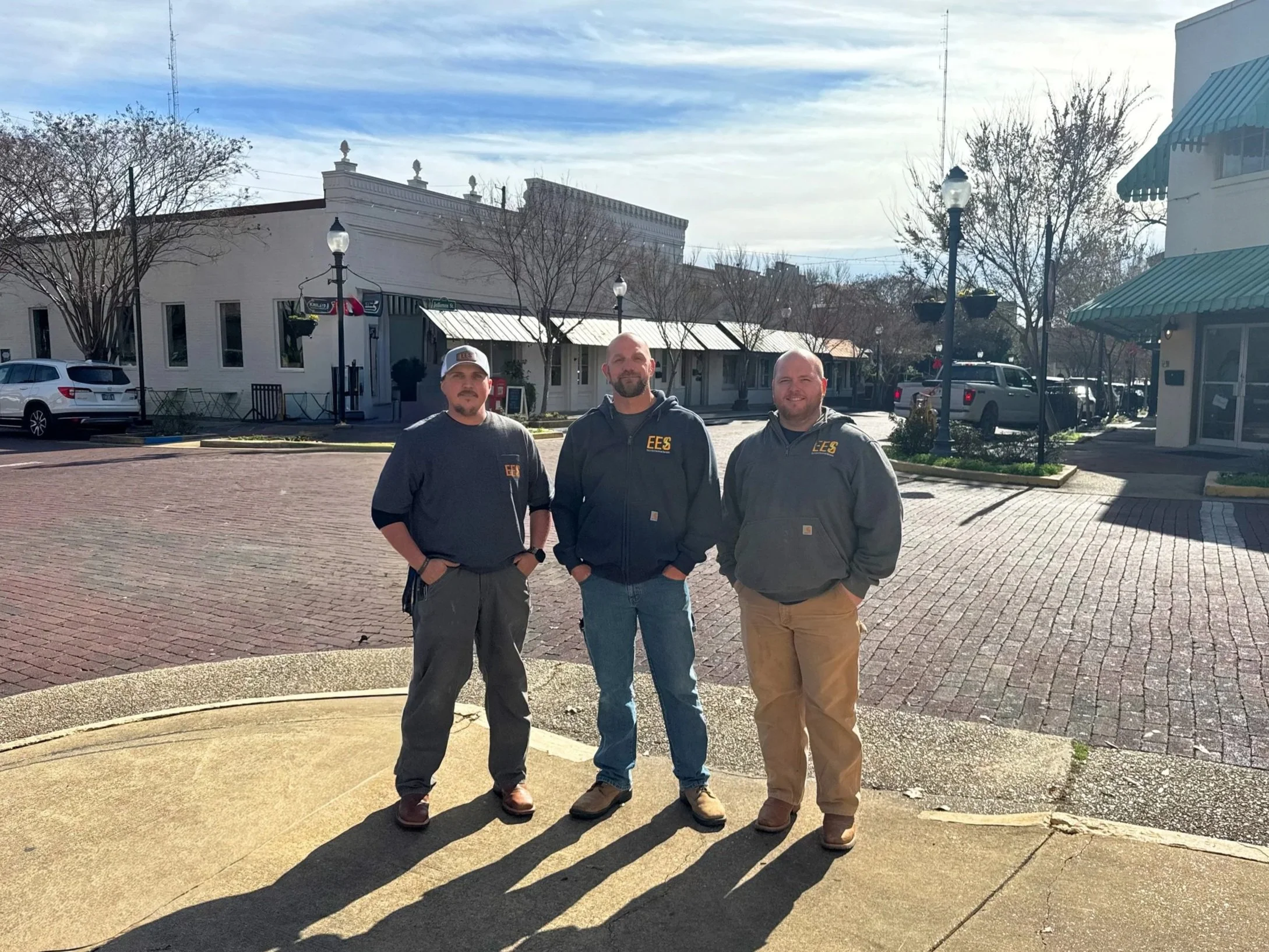 Three men standing on a brick sidewalk in a small town, with shadows cast on the ground during daytime, wearing dark jackets with "EES" logos, in front of storefronts and parked cars.