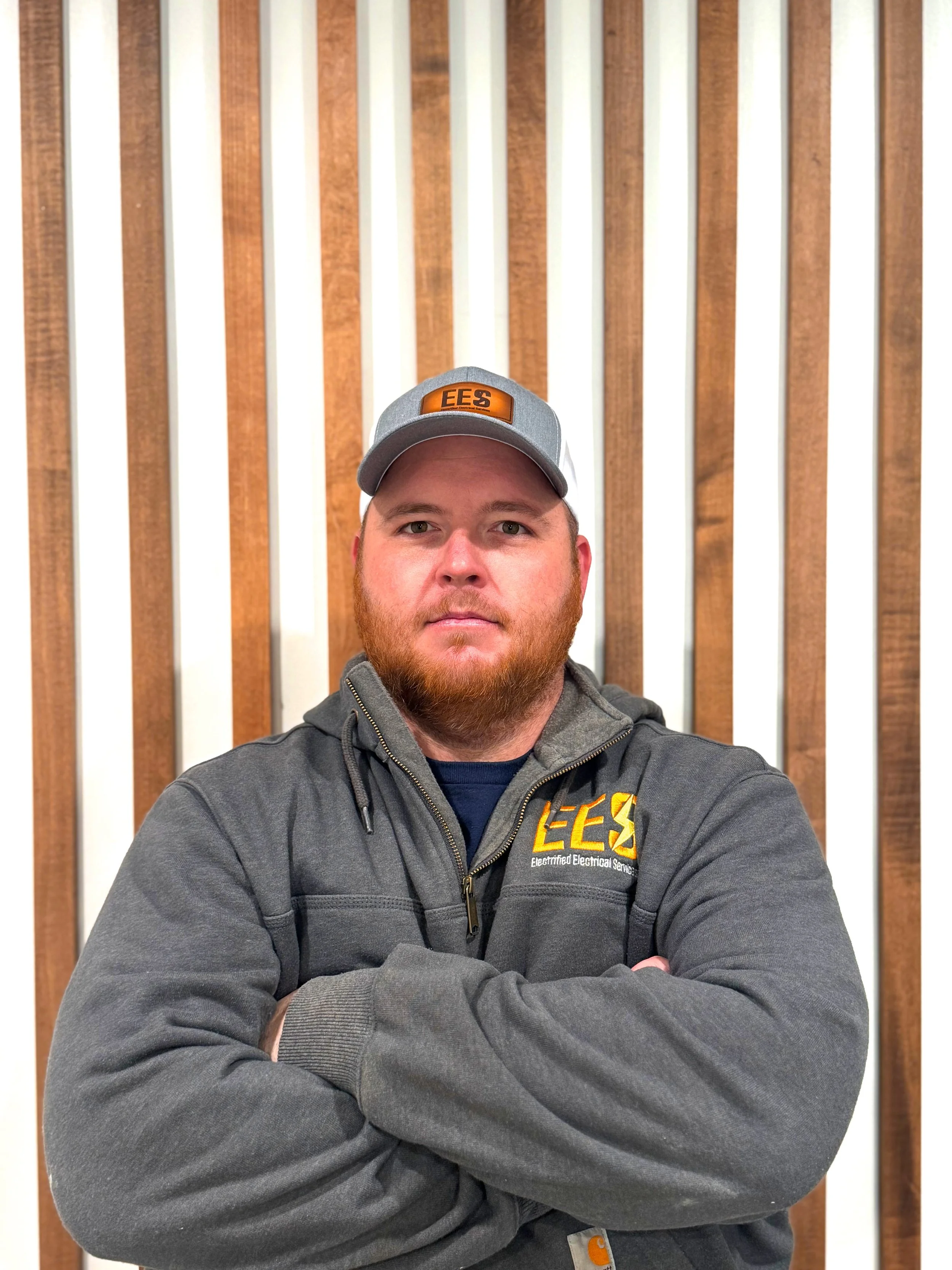 A man with a beard and mustache wearing a gray baseball cap with the logo 'EES' and a gray work jacket with the same logo, standing with arms crossed in front of a wooden and white panel background.