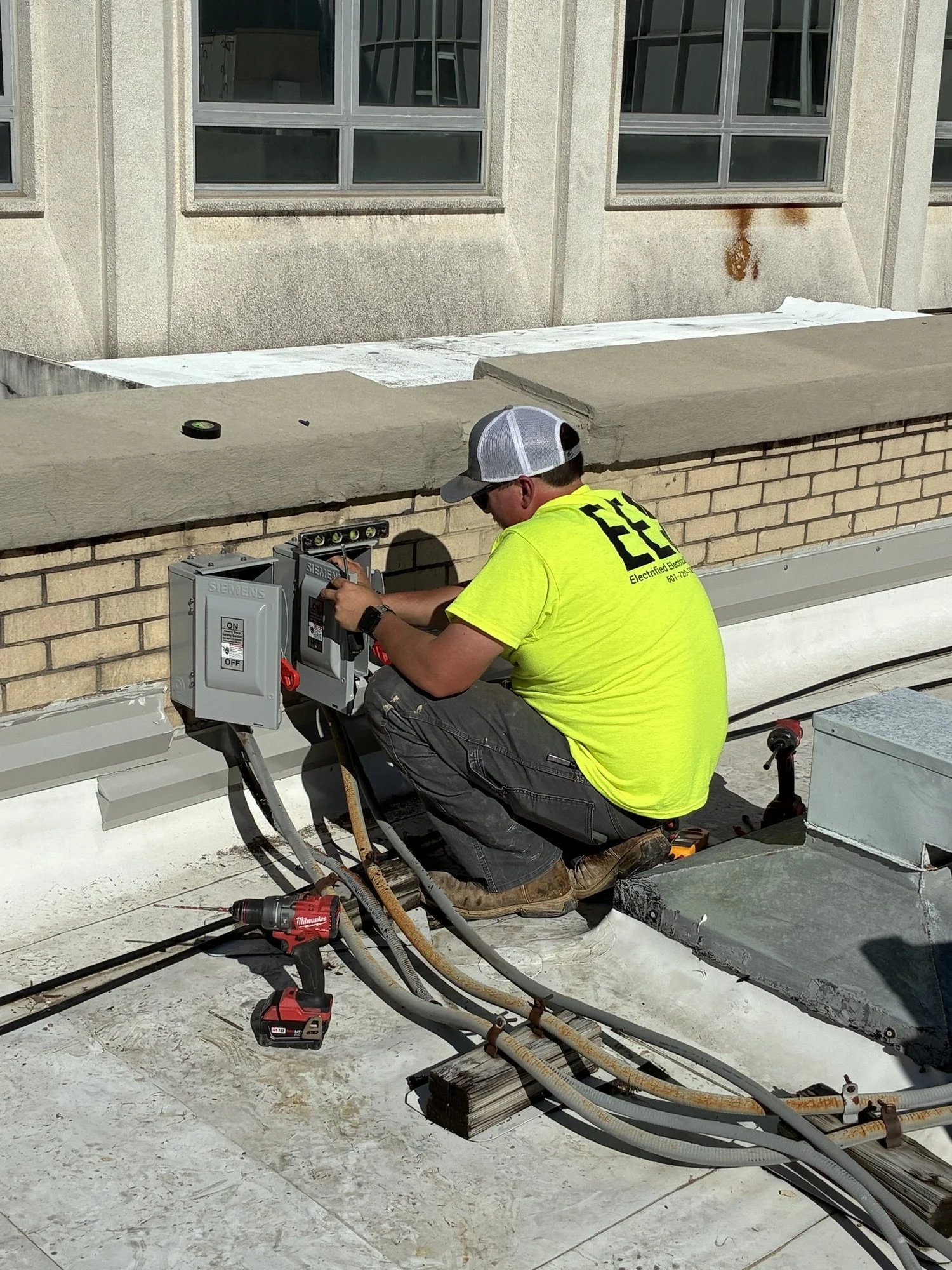 A worker wearing a yellow shirt, gray shorts, and a white and gray cap is kneeling on a rooftop, working on electrical or HVAC equipment. The worker's shirt has the letters 'EE' on the back, and there are tools, cables, and electrical boxes around hi