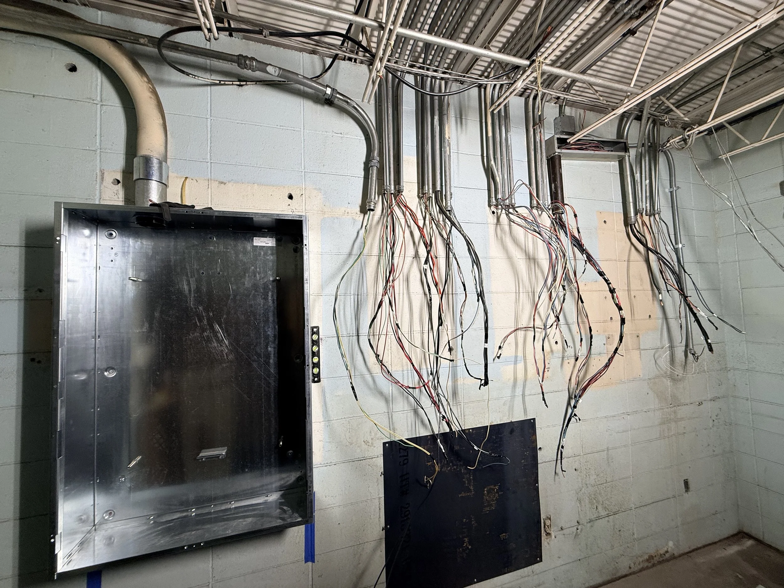 Electrical wiring and conduit pipes exposed on the wall of an unfinished or under renovation room, with an empty metal electrical box and a black panel or cover on the wall.